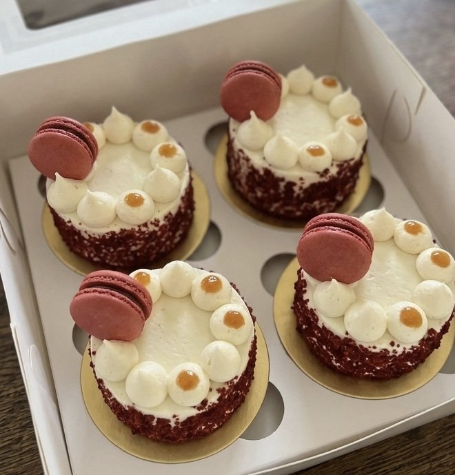 Four decorated mini cakes in a white box, each with a raspberry macaron, cream dollops around the edge, and a caramel topping.
