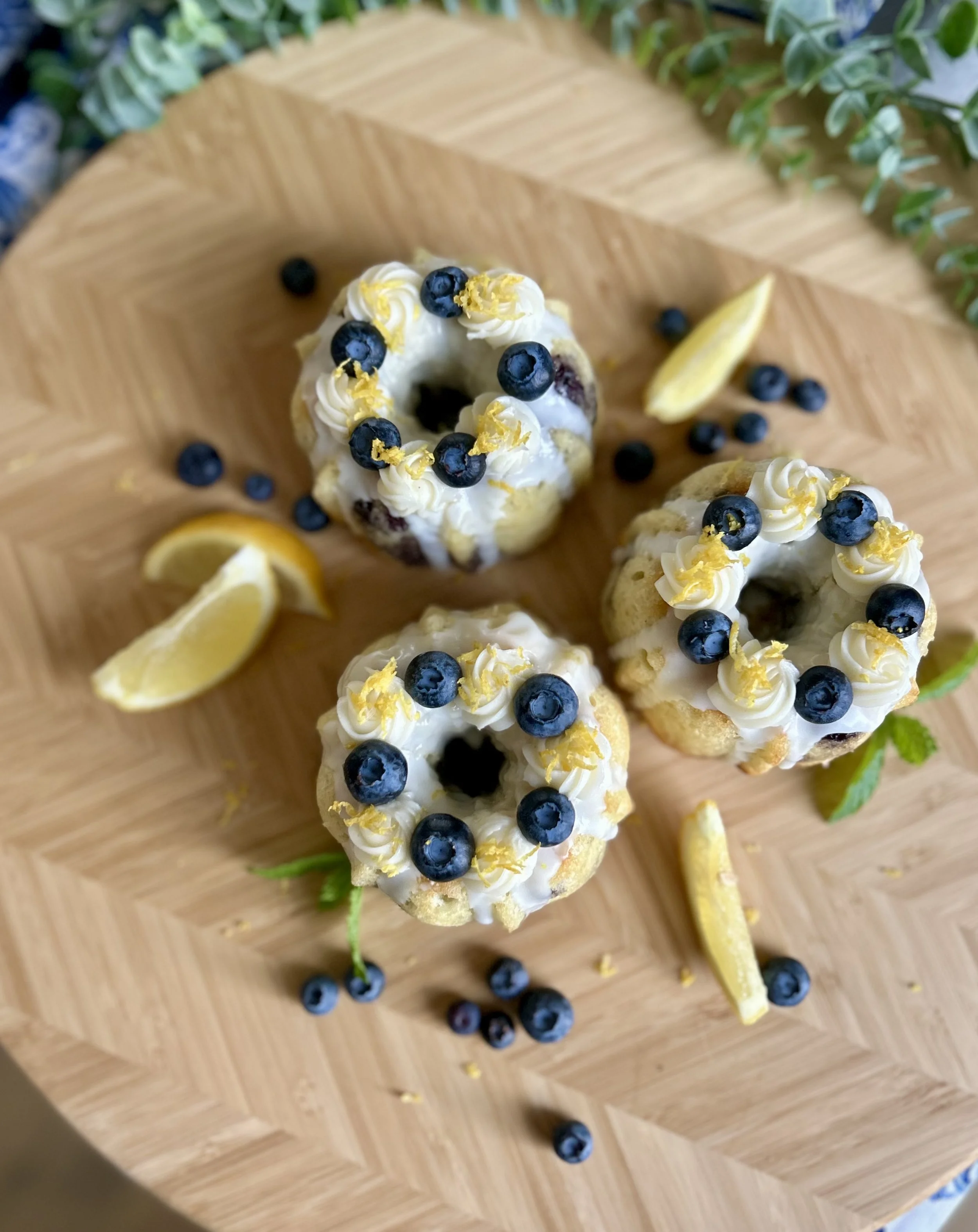 A wooden cutting board with lemon slices, blueberries, and small cakes topped with blueberries, lemon zest, and white cream.