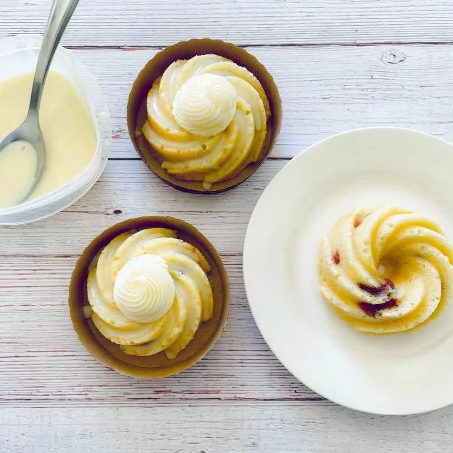 Two lemon tarts with whipped topping on top and one uncut lemon tart on a white plate, with a bowl of lemon curd with a spoon on a white wooden table.