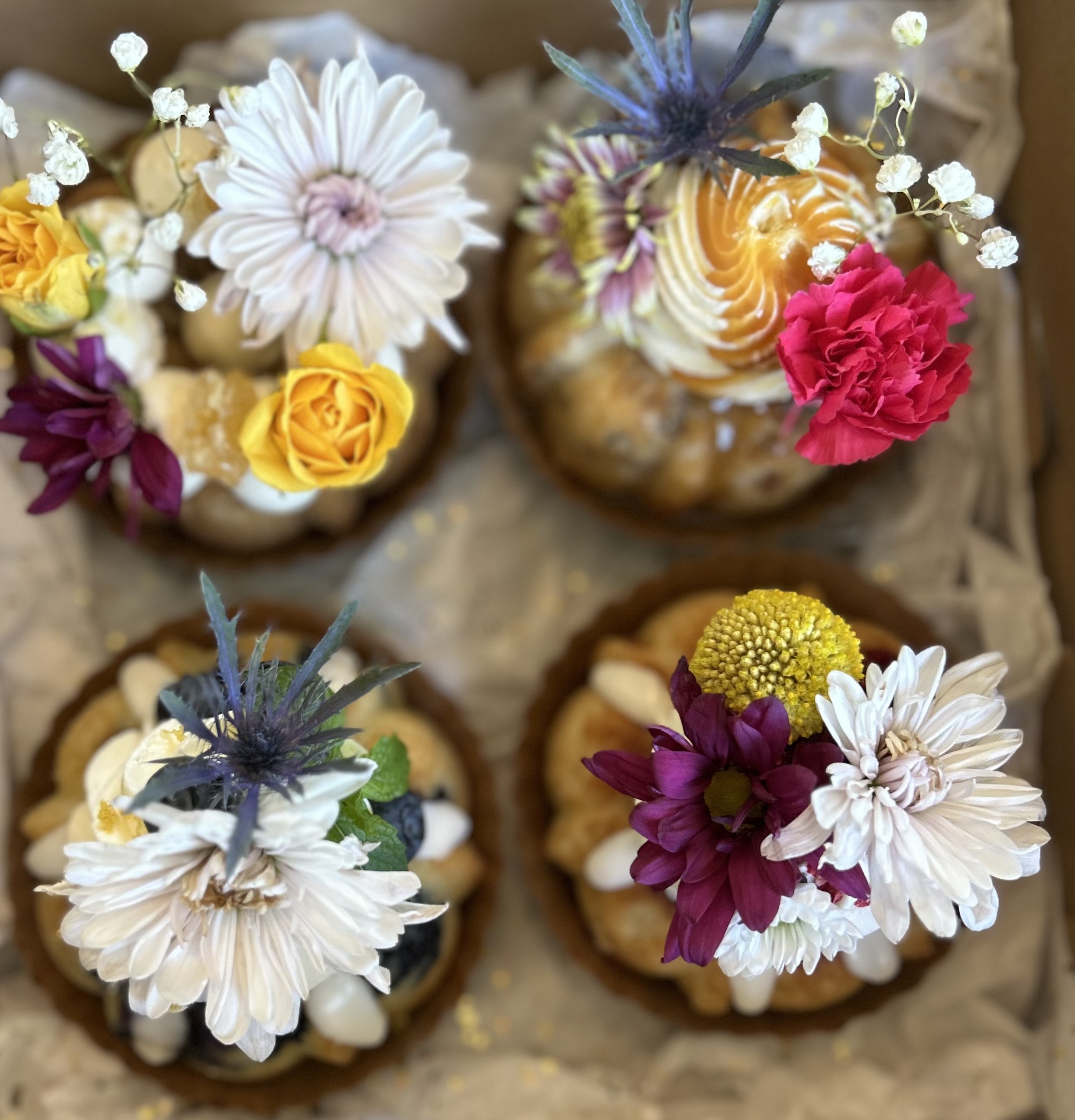 Close-up of a bouquet of various colorful flowers, including white, yellow, pink, and purple blossoms, with some tiny white buds and green leaves visible.