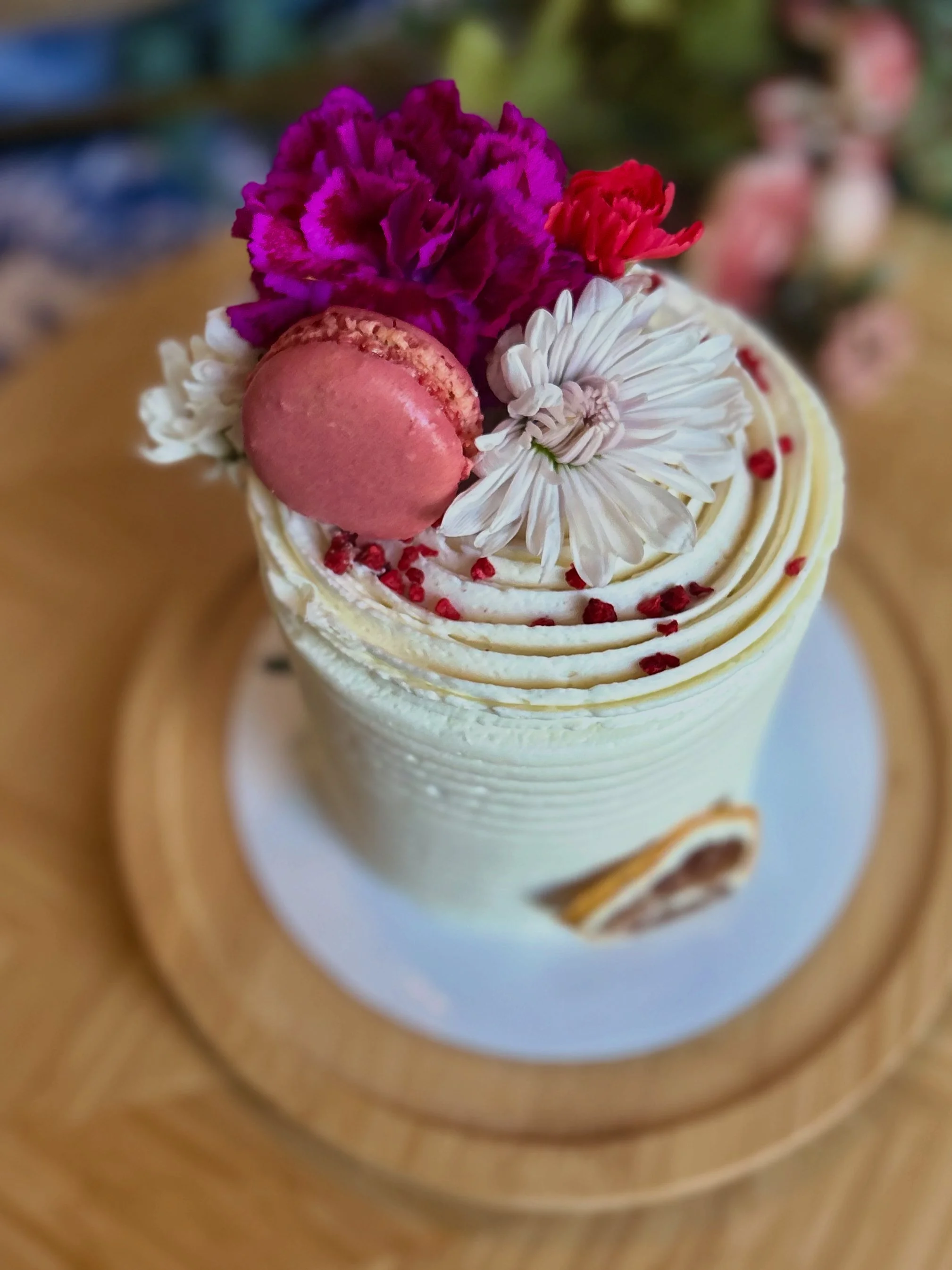 Close-up of a decorated cupcake with white frosting, topped with a pink macaron, purple and red flowers, and red sprinkles, on a white plate with a wooden rim.