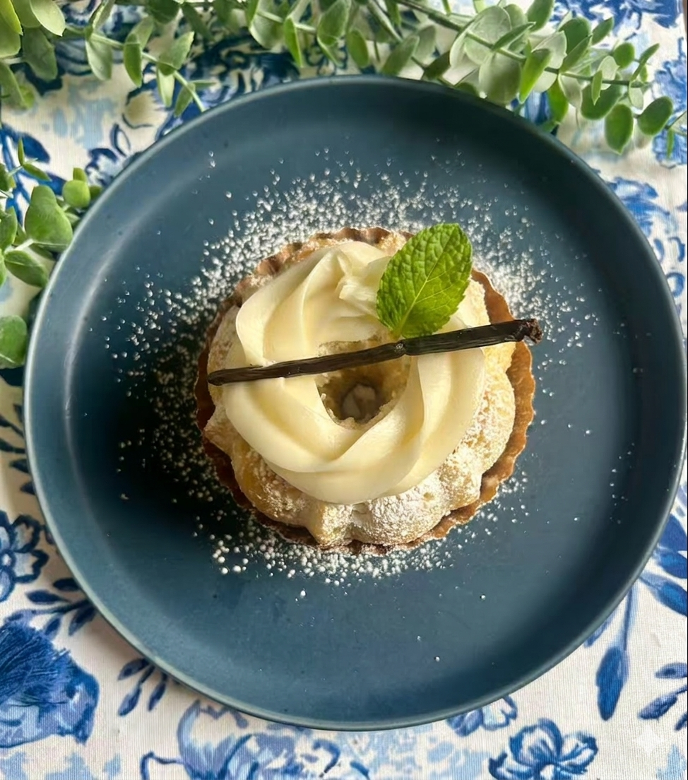 A dessert with white frosting, a powdered sugar dusting, a vanilla bean, a mint leaf, and a decorative blue plate, on a floral tablecloth with greenery.