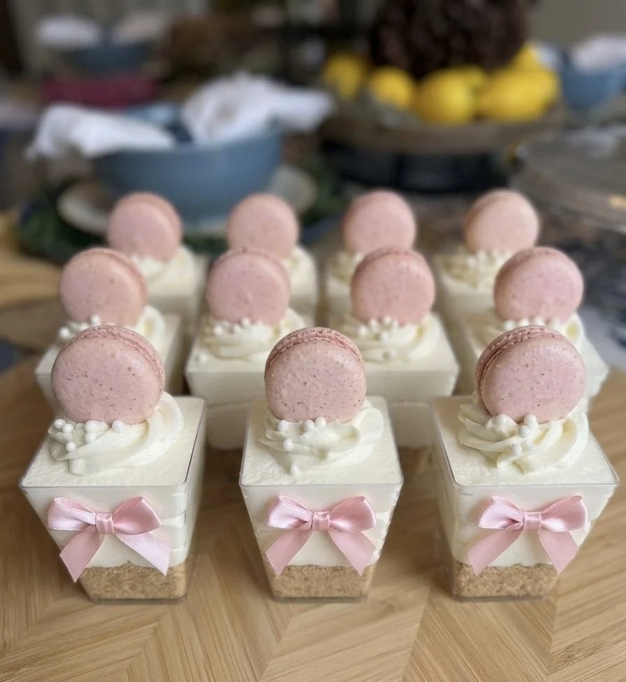 Small layered desserts in clear cups with white cream, pink macarons, and pink bows on the cups, arranged on a wooden surface.