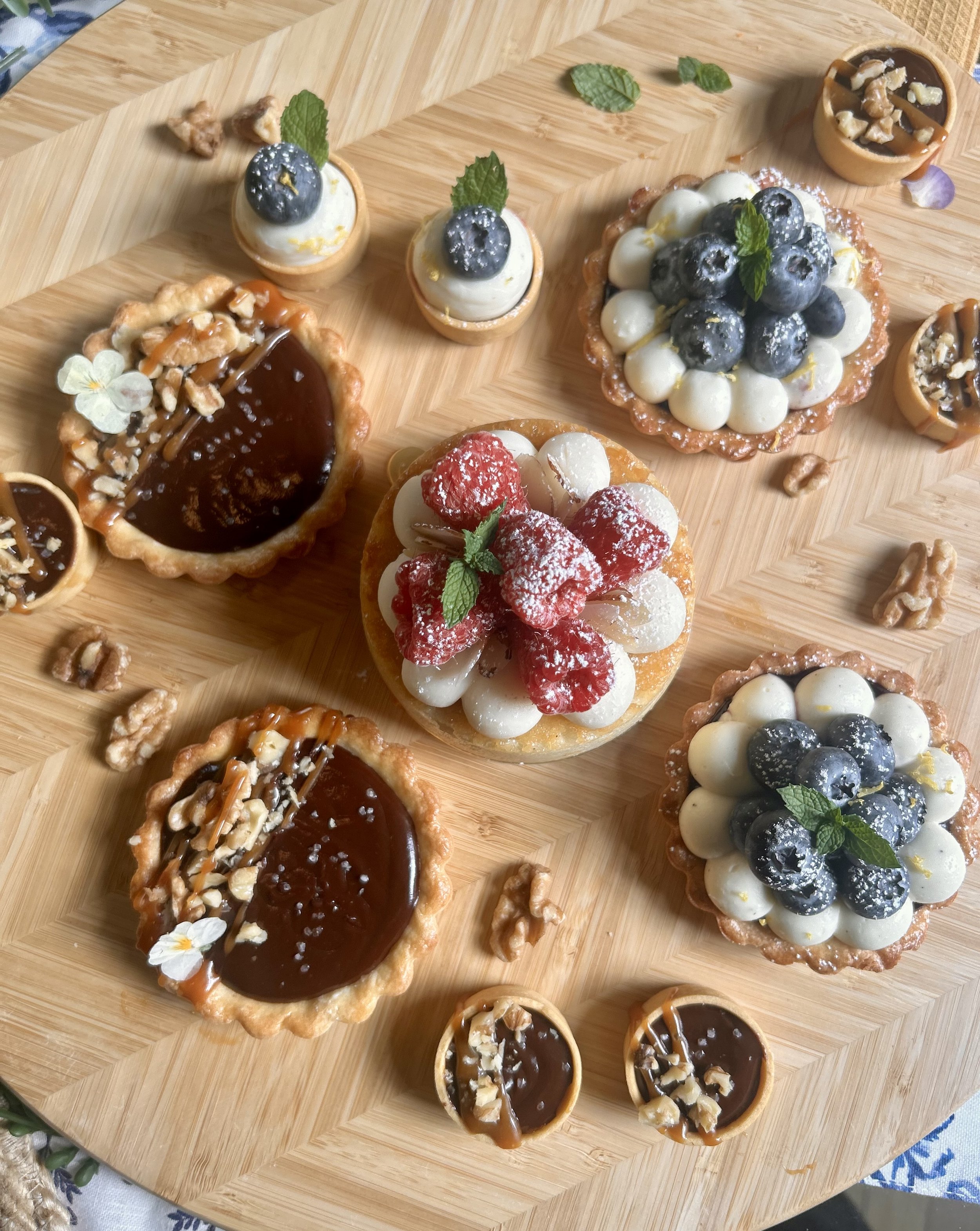 Assorted mini pies with various toppings including blueberries, strawberries, nuts, and chocolate, arranged on a wooden surface.
