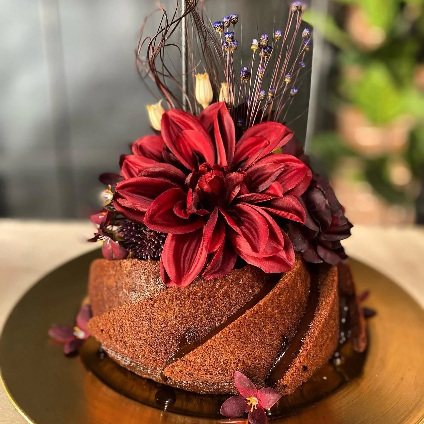 A close-up of a chocolate cake topped with red and purple flowers, drizzled with chocolate sauce on a gold plate.