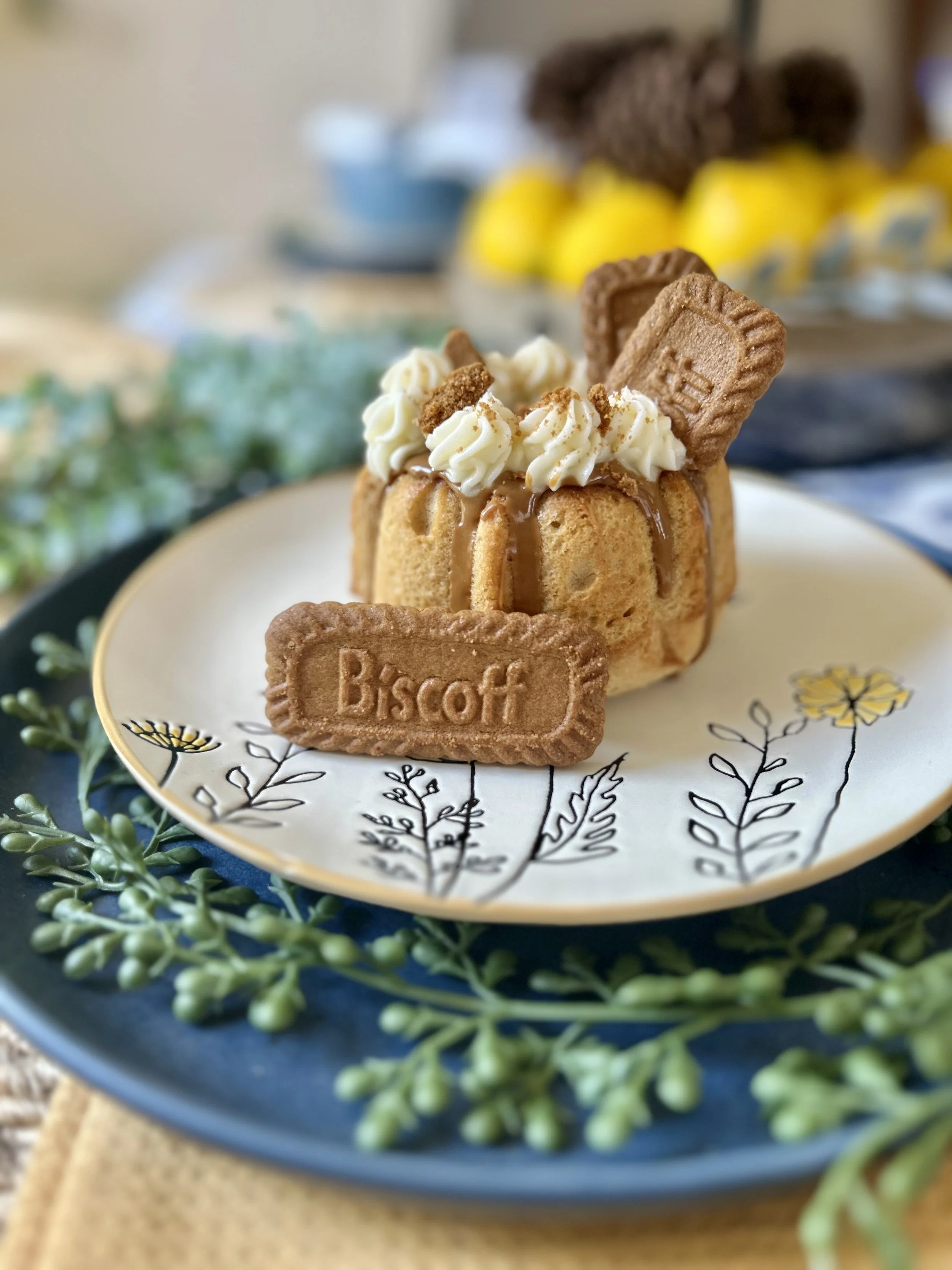 A small dessert with a biscuit base, topped with whipped cream, caramel sauce, and two biscuits, presented on a plate with a floral design, with a blurred background of teacups and yellow decorations.