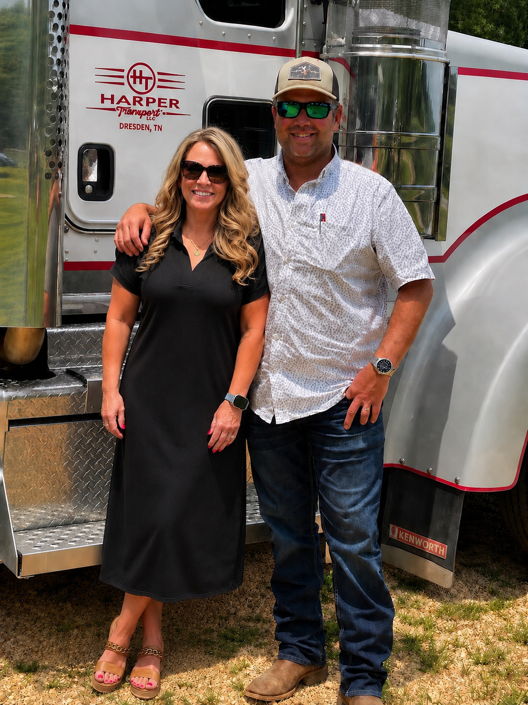 A man and woman standing in front of a large white and silver truck. The woman is wearing a black dress and sunglasses, and the man is wearing a white patterned shirt, jeans, sunglasses, and a cap. They are smiling, with the man's arm around the woman's shoulder.