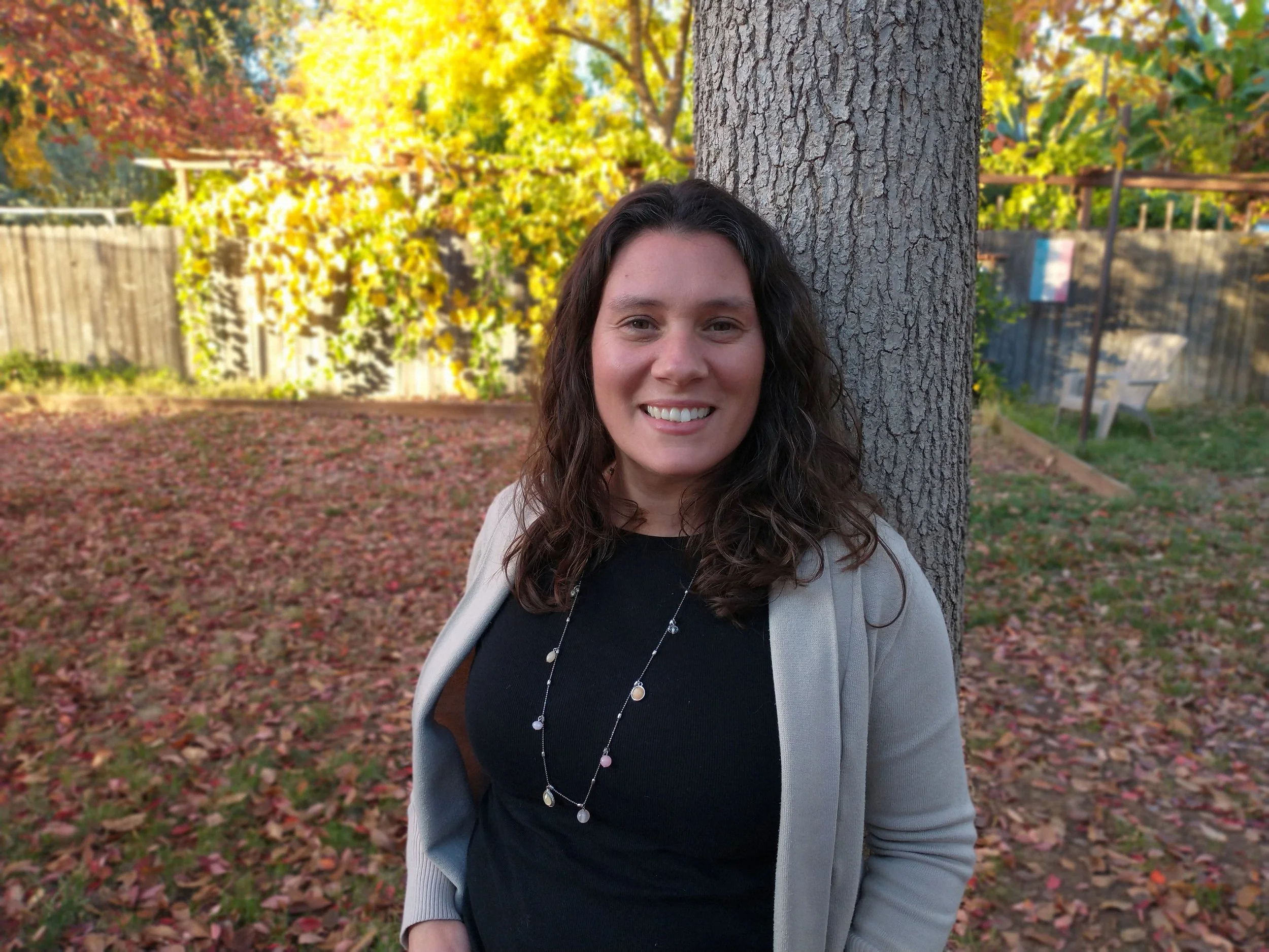 A woman with long curly hair smiling outdoors in autumn, standing beside a large tree with yellow and red foliage in the background.