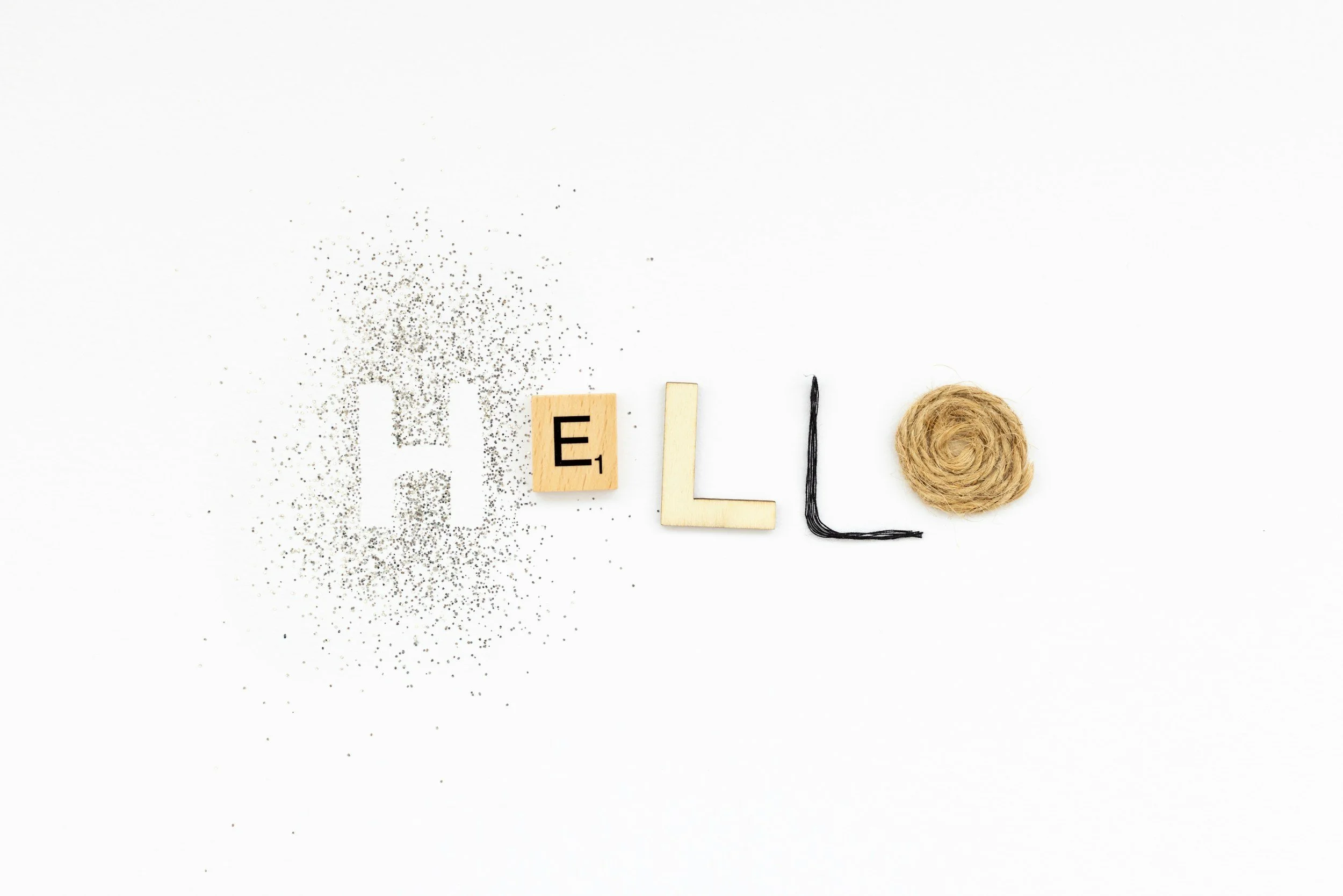 Letters spelling out the word 'HELLO' on a white background, with one letter made of black glitter, and the letter 'E' on a Scrabble tile, a piece of twine on a spool, and scattered black glitter.
