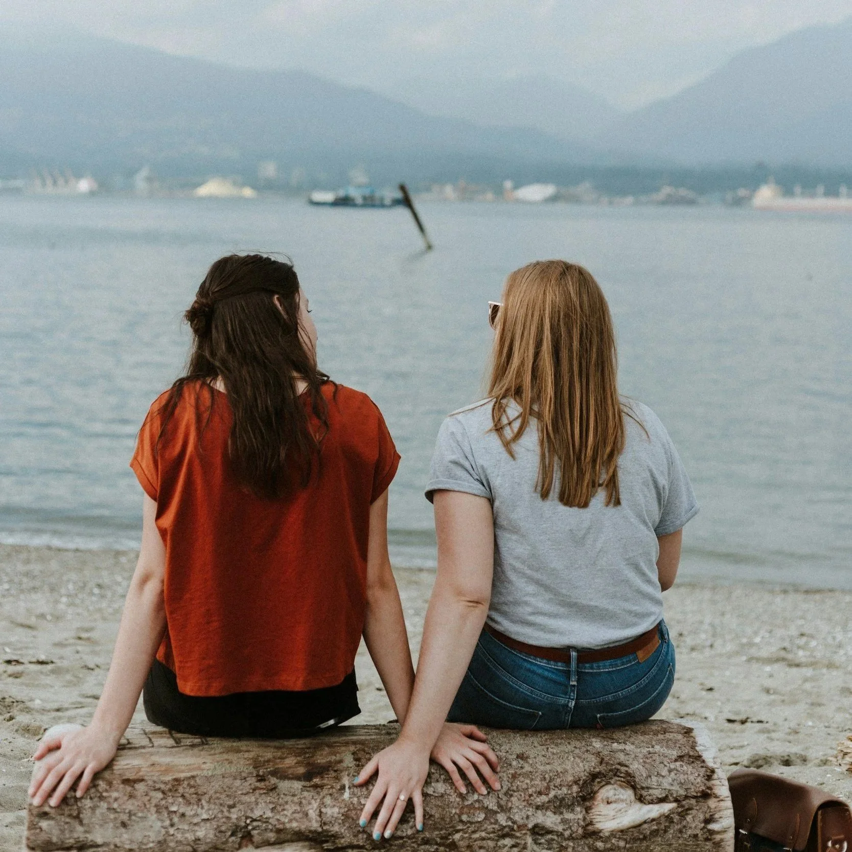 Two women sitting on a log at the beach, holding hands, facing the water with ships and mountains in the background.