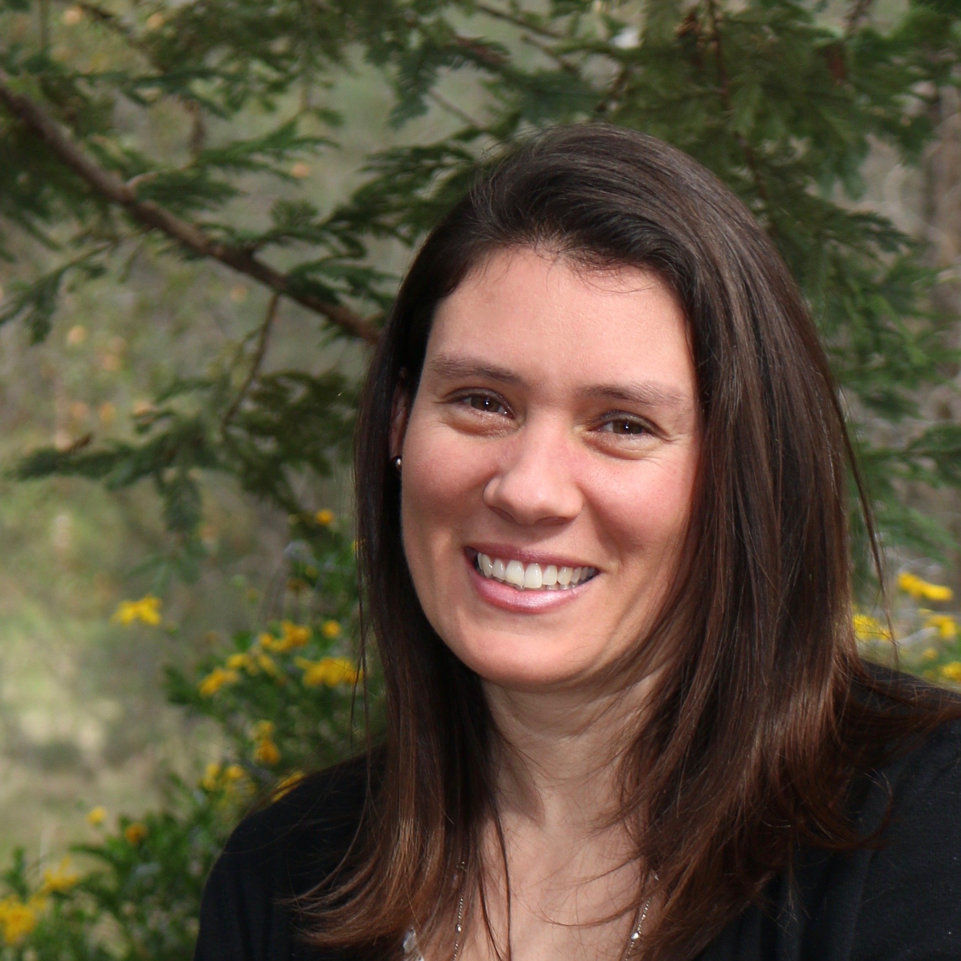 The photo shows the smiling face of a woman with long brown hair, set against a backdrop of green foliage.