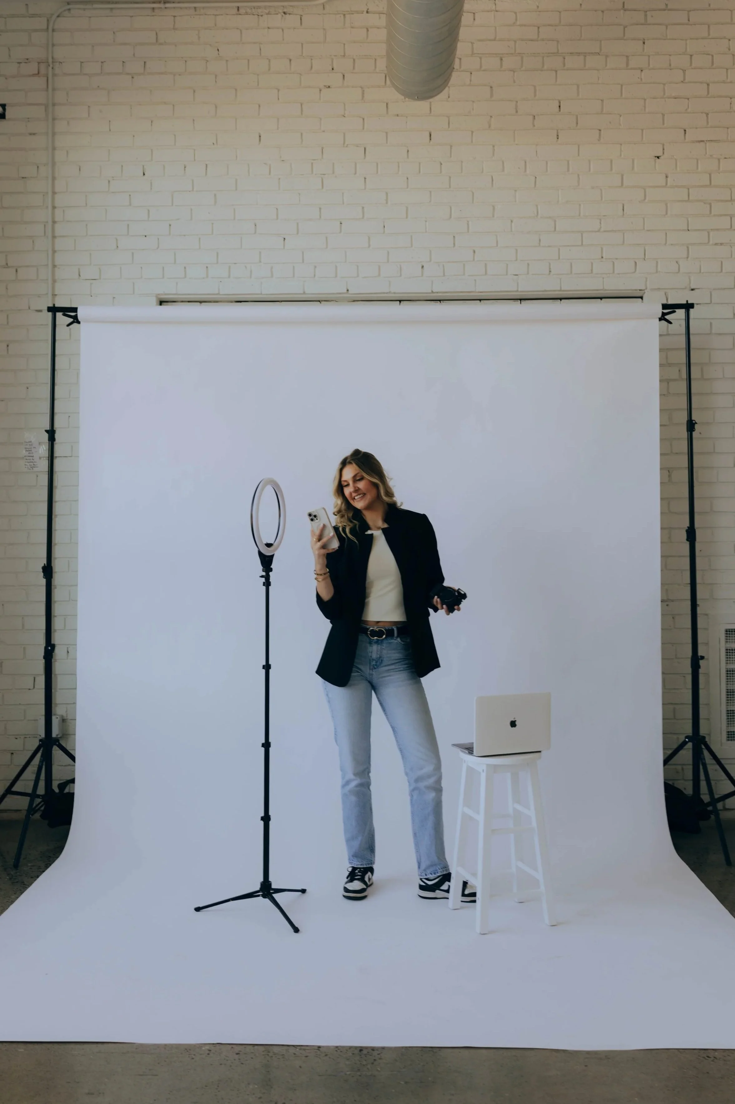A woman with blonde hair, wearing a black blazer, white shirt, and blue jeans, is standing in a photo studio with a white backdrop. She is smiling and looking at her phone, holding a camera in one hand. There is a ring light on a stand in front of her, and a laptop on a small white stool nearby. The studio has a brick wall and a ventilation pipe on the ceiling.