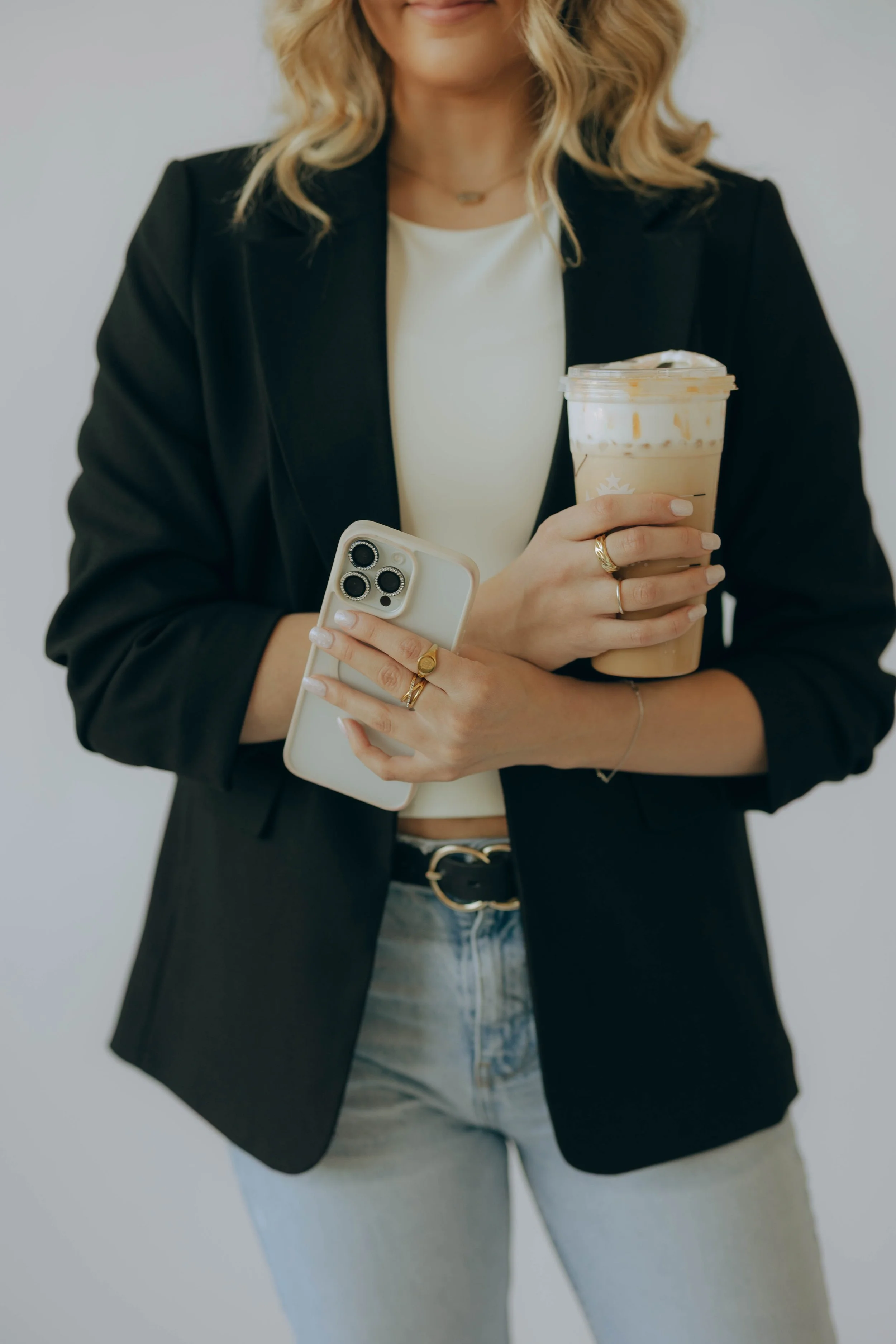 A woman in a black blazer and light jeans holding a Starbucks iced coffee and a smartphone, with her mouth slightly smiling at the camera.