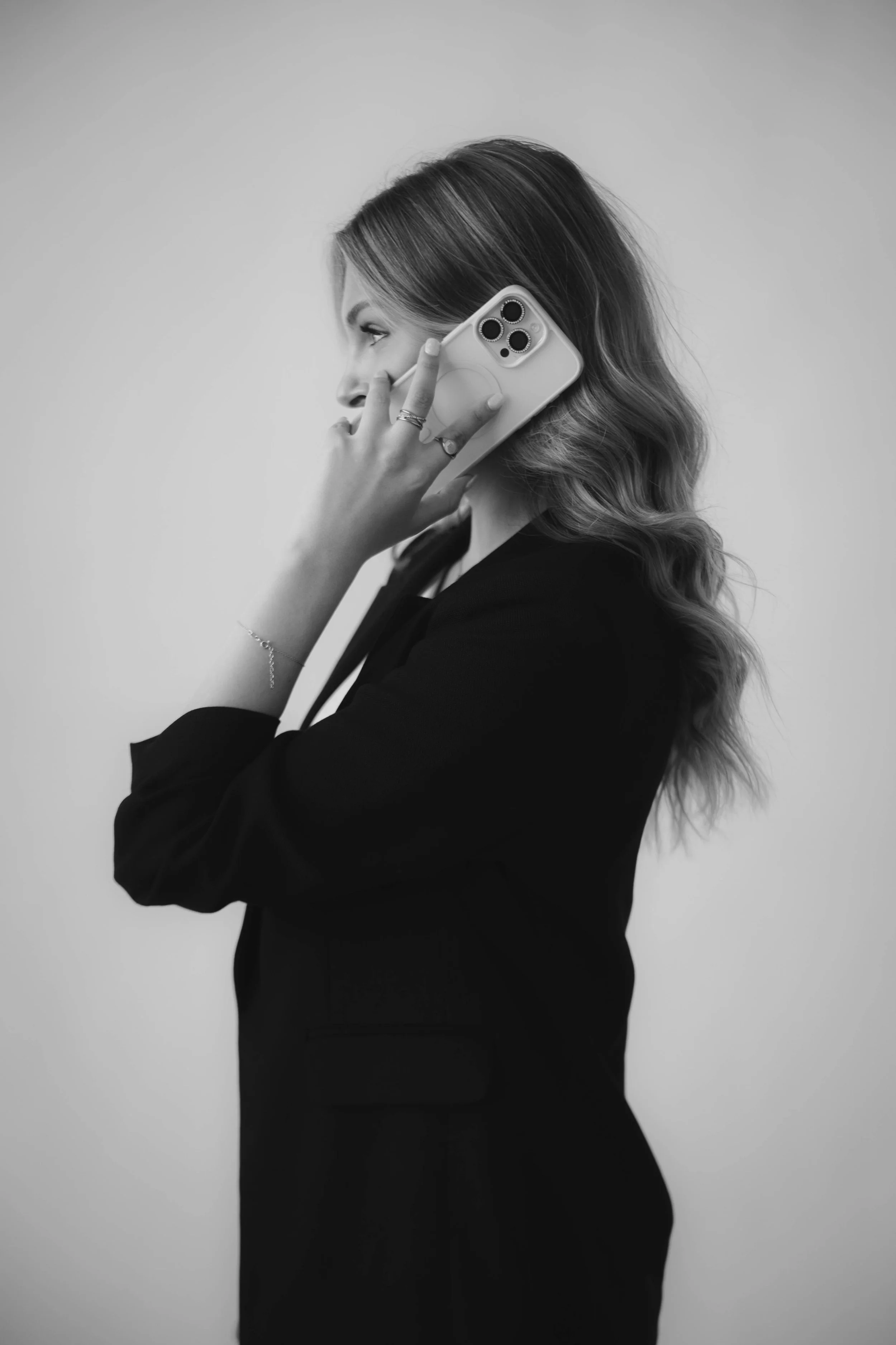 A woman with shoulder-length wavy hair, dressed in a black blazer, is talking on her smartphone with a plain light-colored wall in the background.