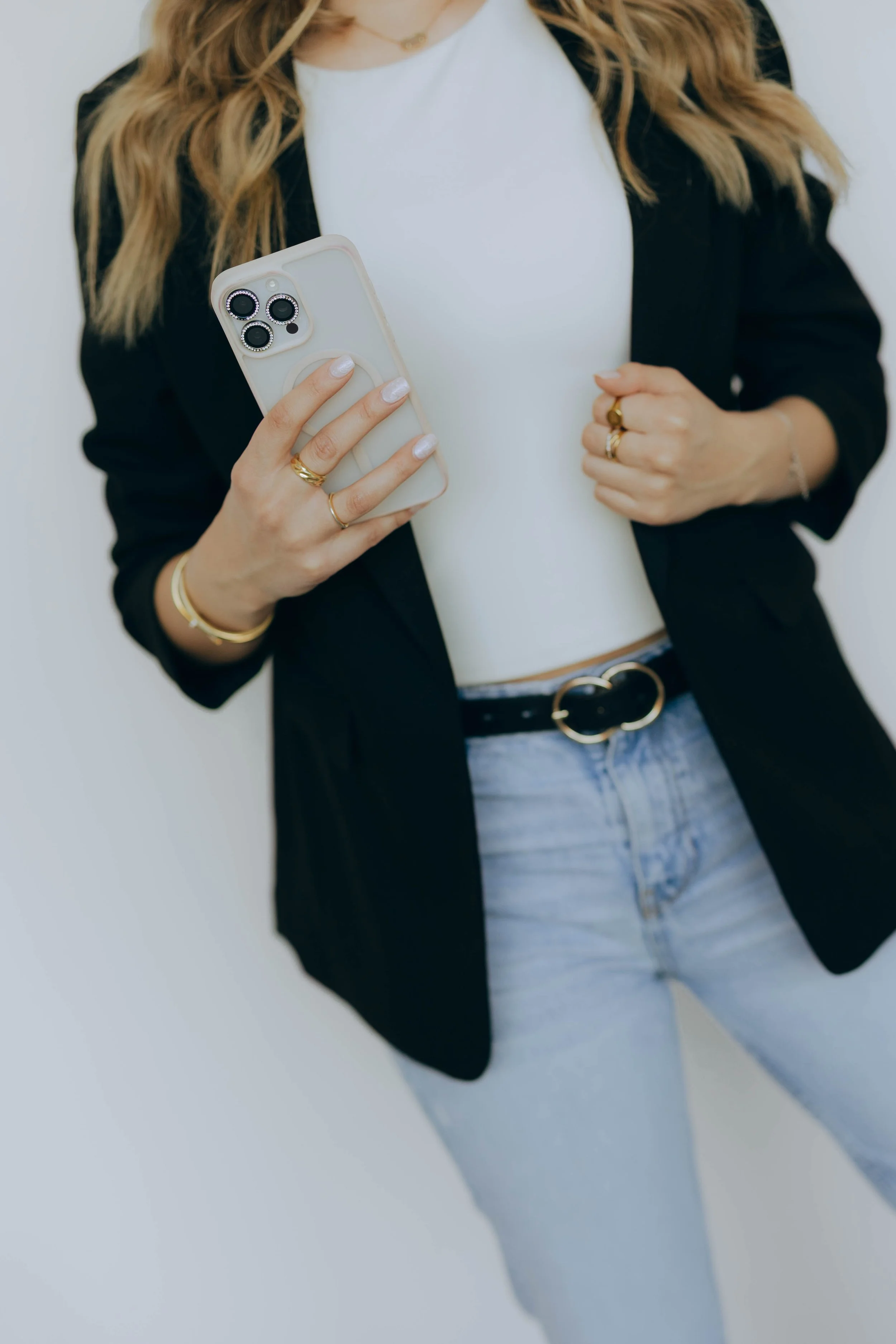 A woman wearing a black blazer, white top, and light blue jeans is holding a smartphone with a white case. She has multiple gold rings and a gold bracelet, and her long, wavy hair is partly visible.