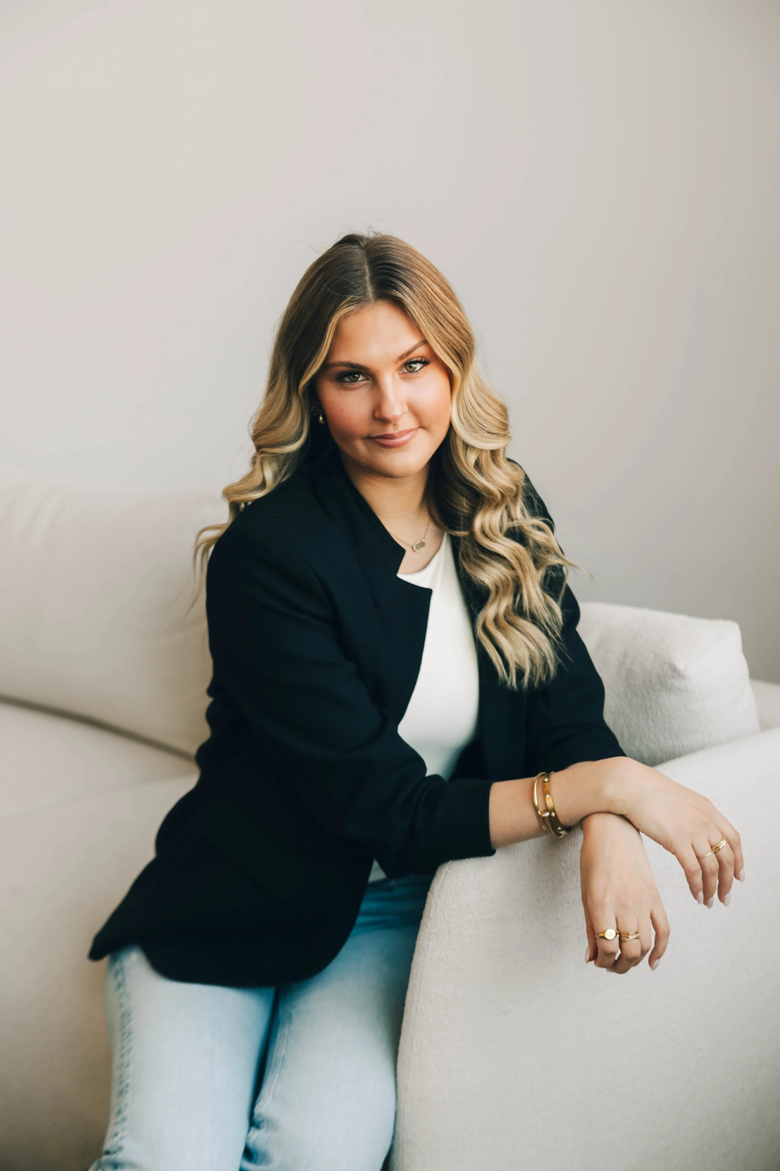 A woman with blonde, curly hair and a black blazer sitting on a beige couch with a relaxed pose, wearing gold jewelry, indoors against a plain wall.