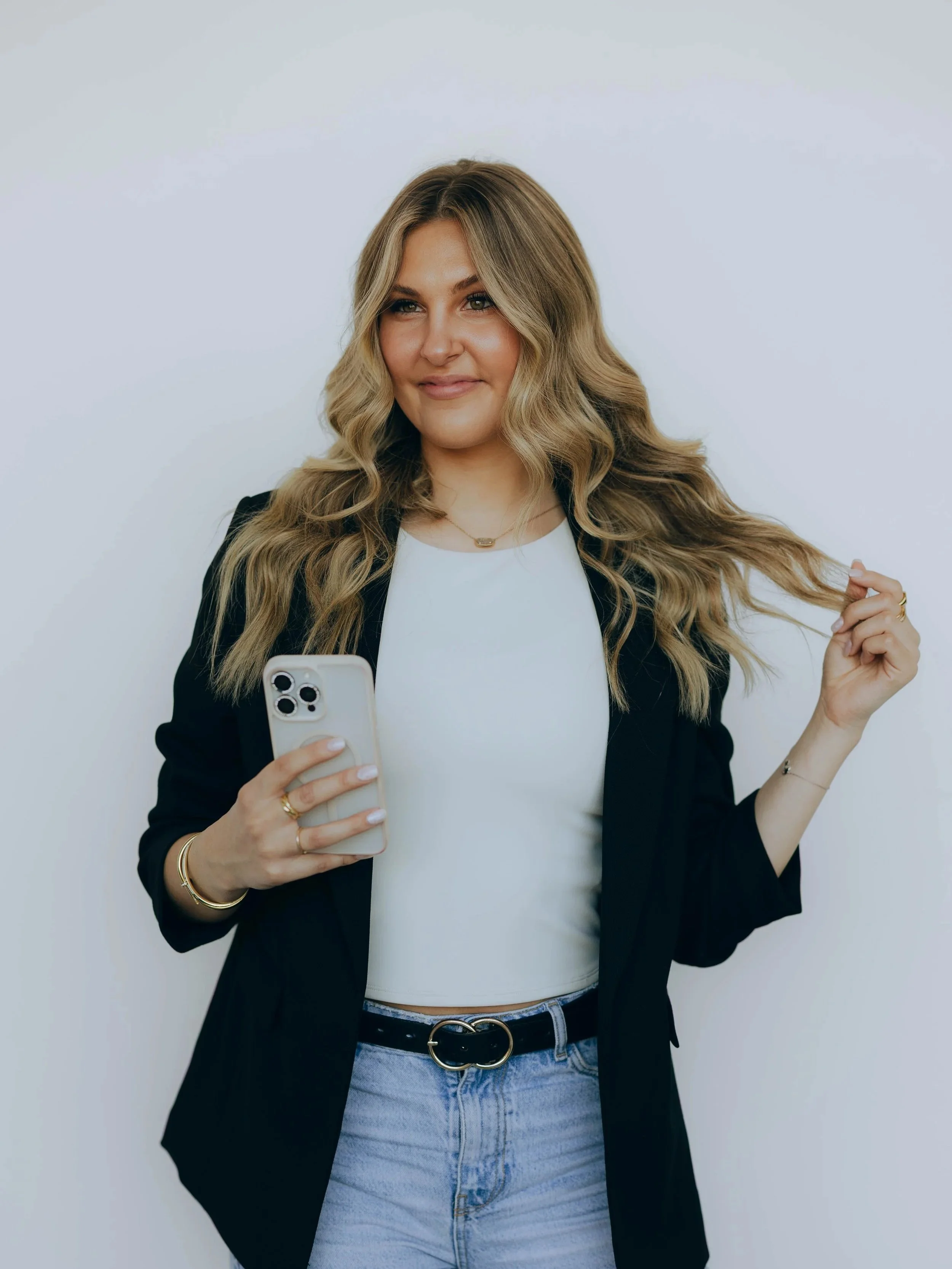 Young woman with long wavy blonde hair wearing a white top, black blazer, and blue jeans, holding a smartphone and standing against a plain white background.