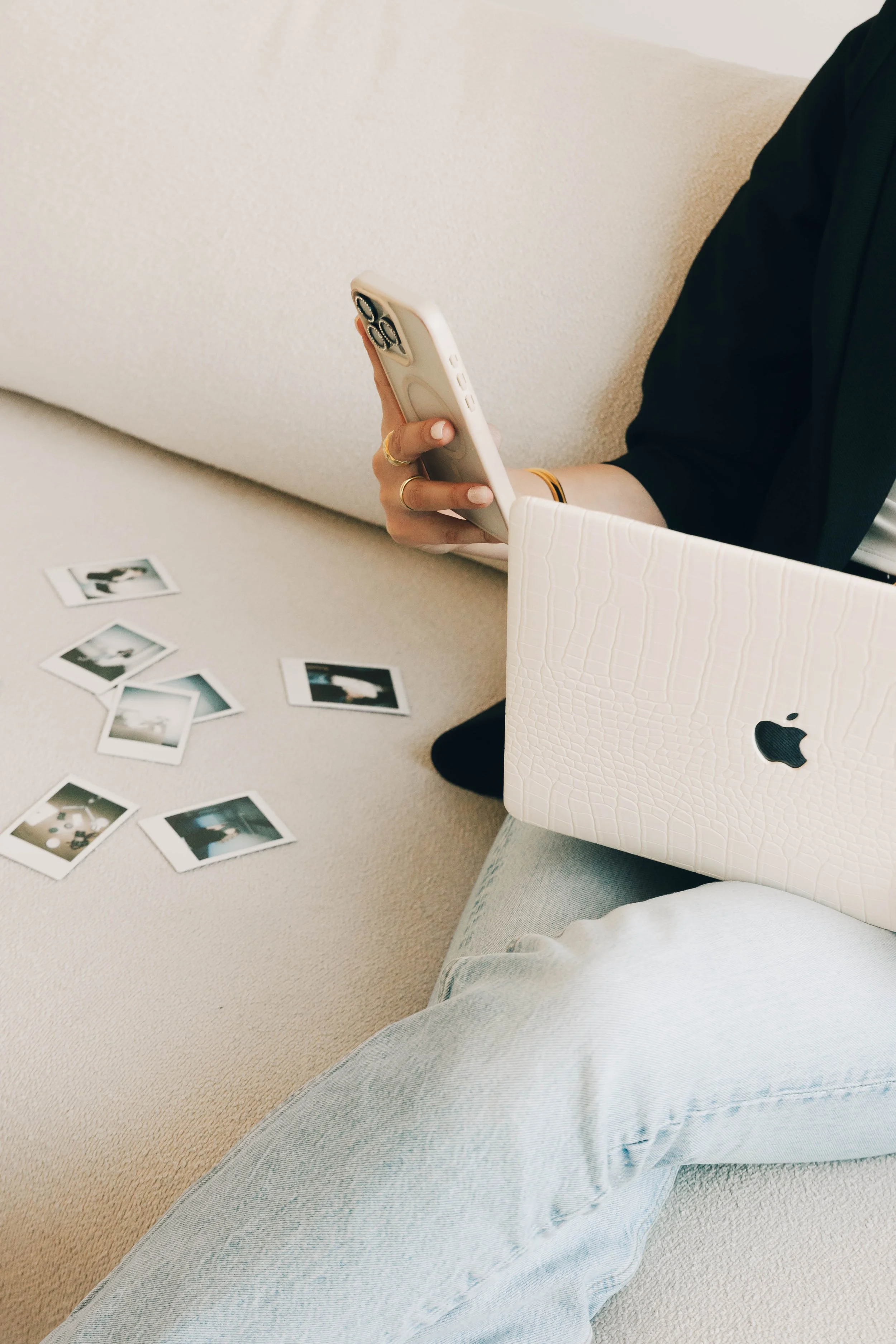 Person sitting on a beige carpeted floor with a white Polaroid camera, several Polaroid photos scattered around, and a closed white Apple laptop on their lap, wearing light-colored jeans and a white shirt.