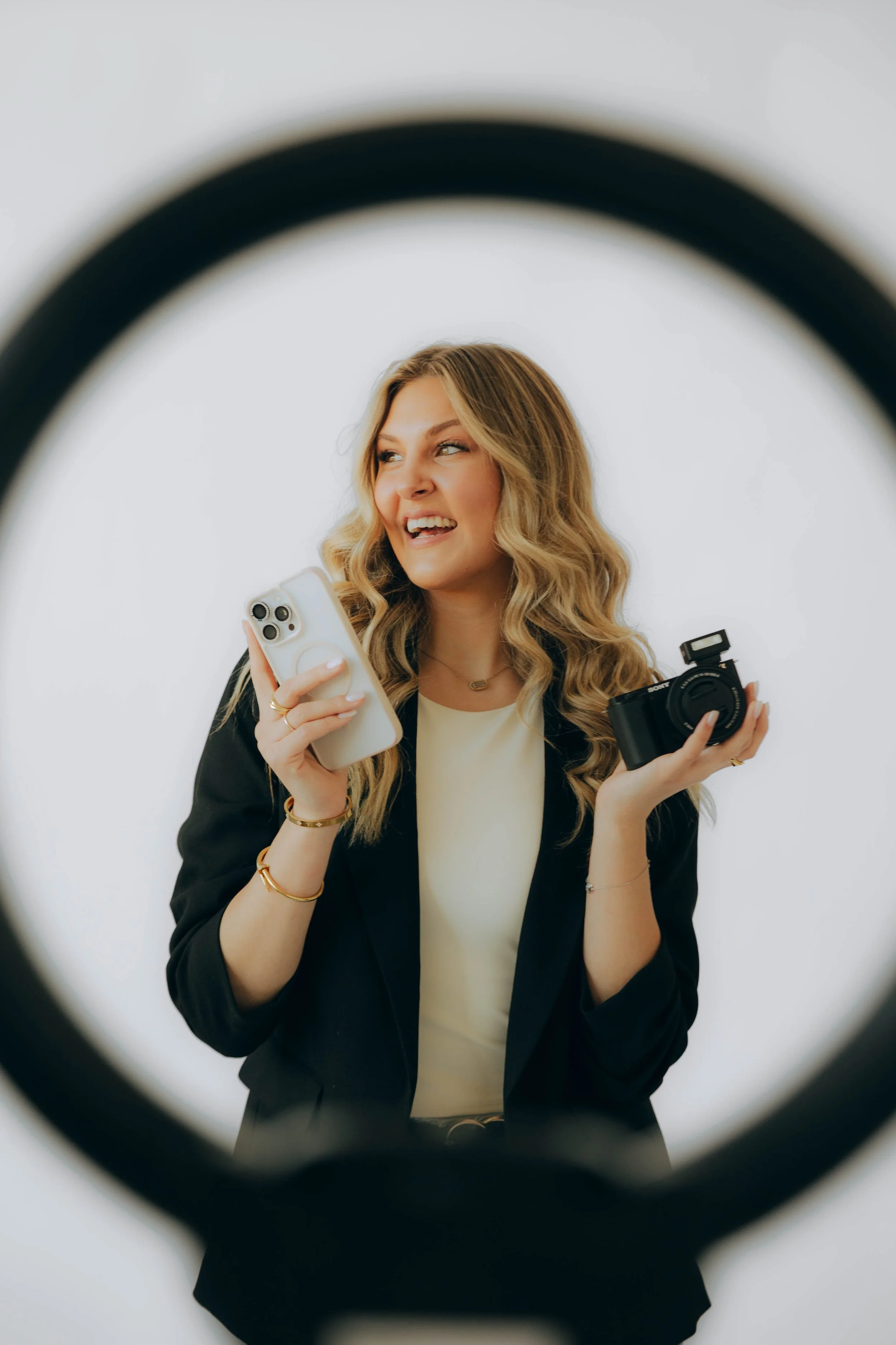 A woman with blonde wavy hair smiling while holding a smartphone and a camera, seen through a circular framing.