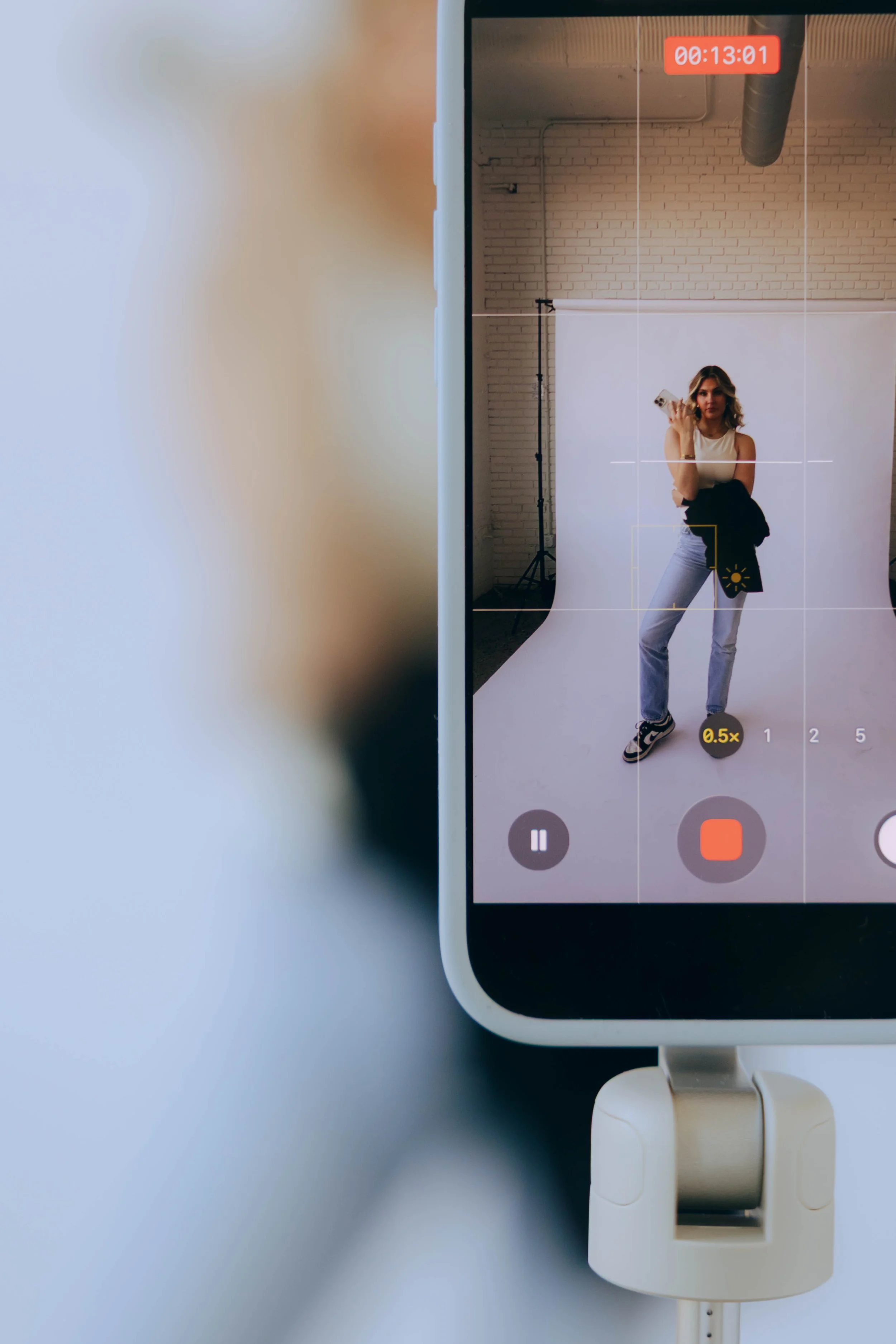 Tablet screen showing a woman taking a selfie in a studio with a white backdrop, holding a phone and a jacket, with a timer at the top and recording controls at the bottom.