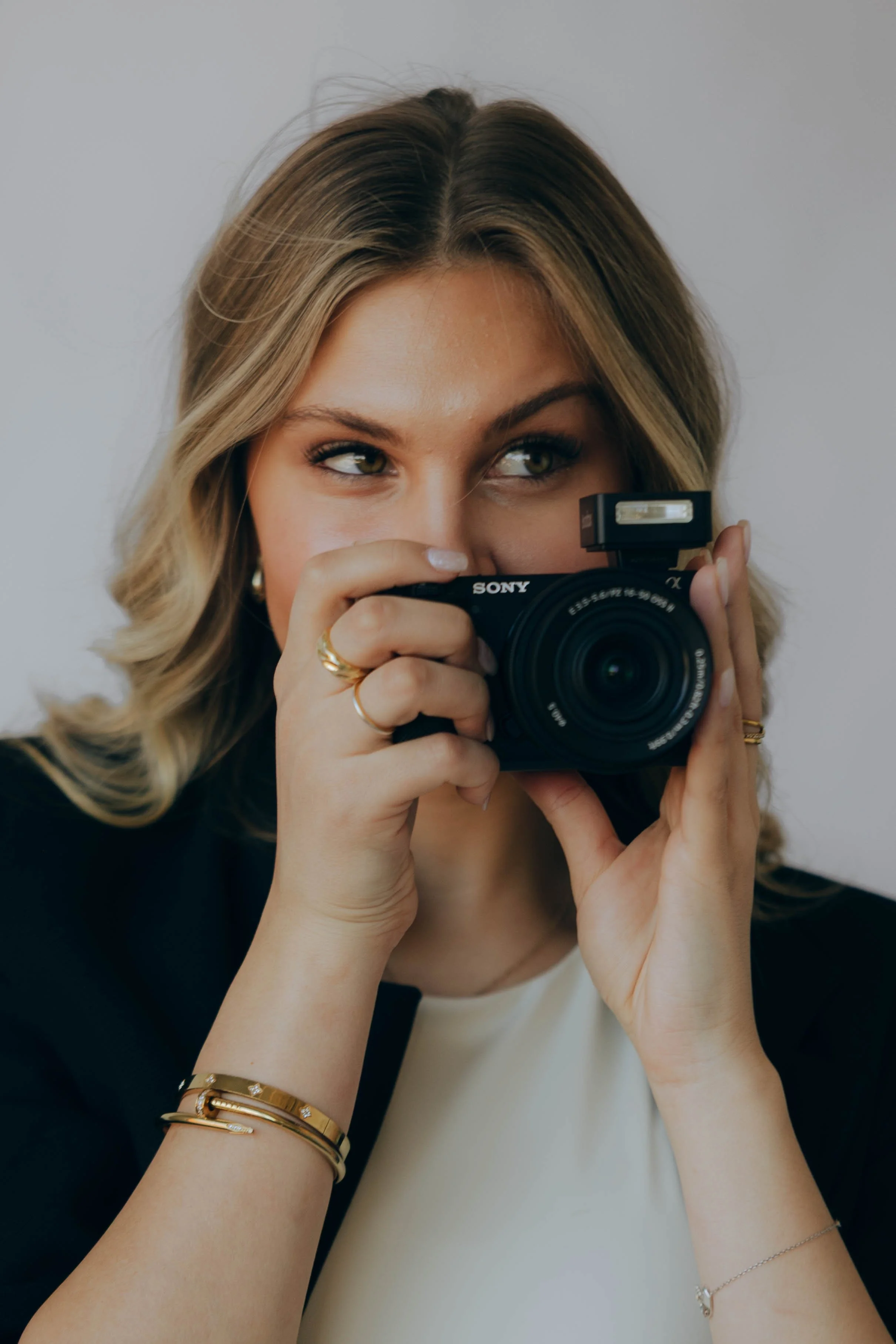 A woman with blonde hair and green eyes is taking a photo with a black Sony camera, holding it up to her face, with a plain light-colored background.