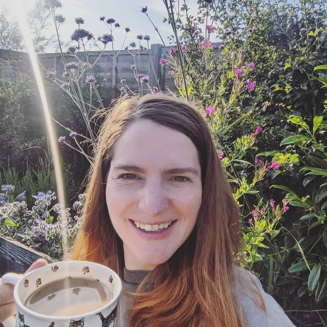 A woman smiling outdoors holding a cup of coffee or tea, with flowers and greenery in the background on a sunny day.