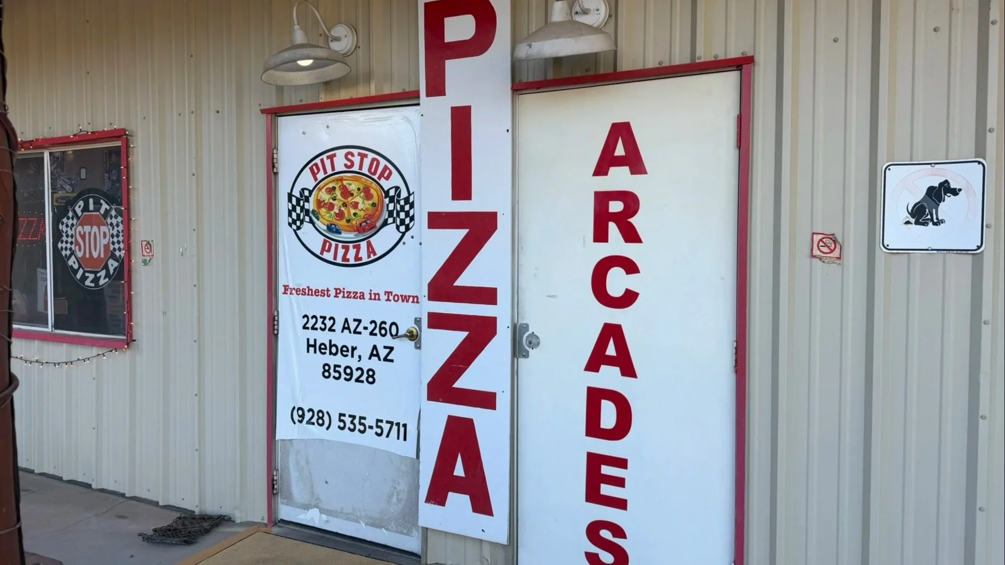 The storefront of a pizza shop called Pit Stop Pizza with a large sign displaying the shop's name, logo with a pizza, and contact details. The building has metal siding, and there is a smaller sign with a dog image and a no-smoking symbol on the wall.