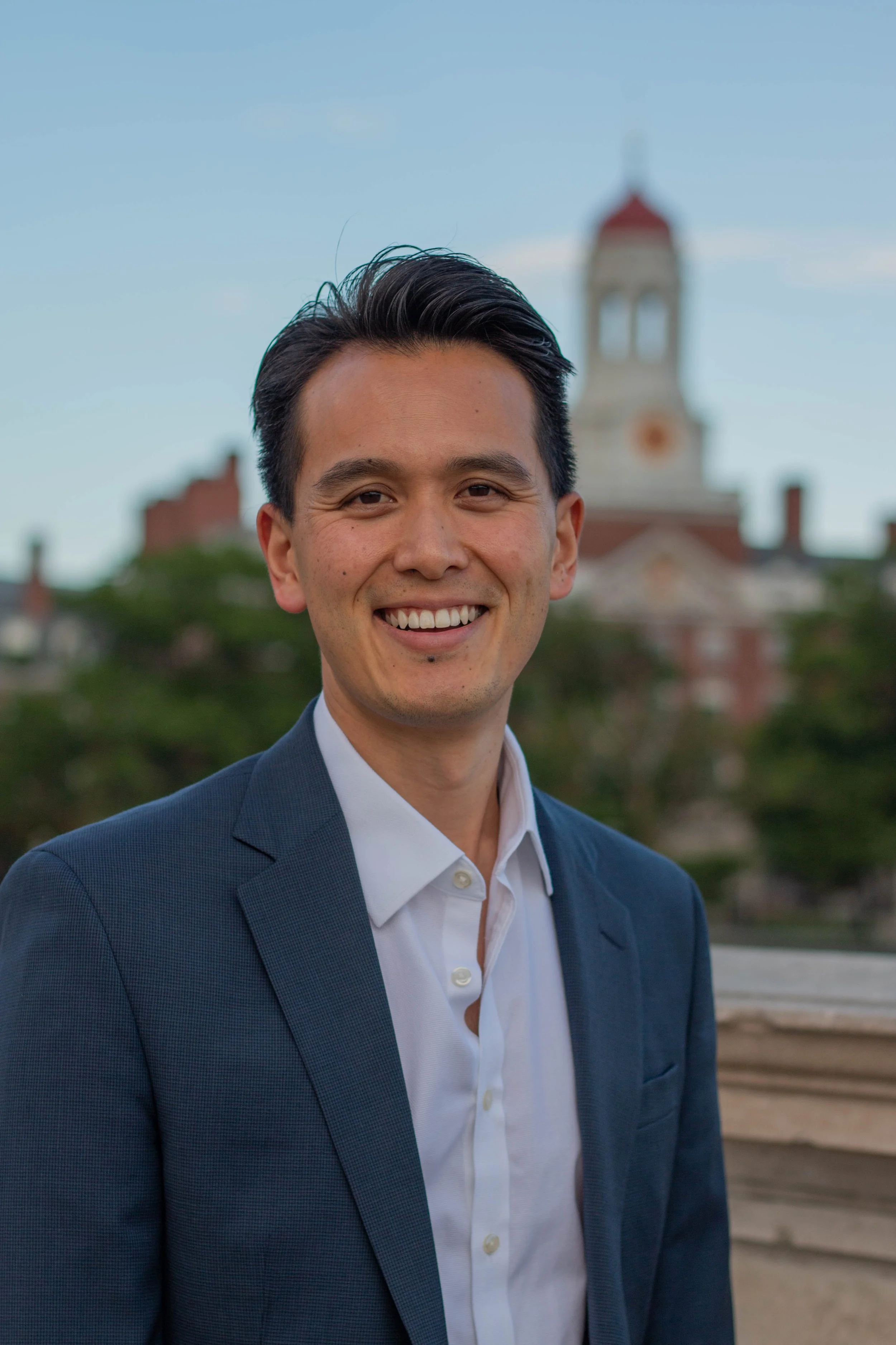 A young man dressed in a dark blue suit jacket and white shirt, smiling outdoors with a historic town hall or clock tower in the background.