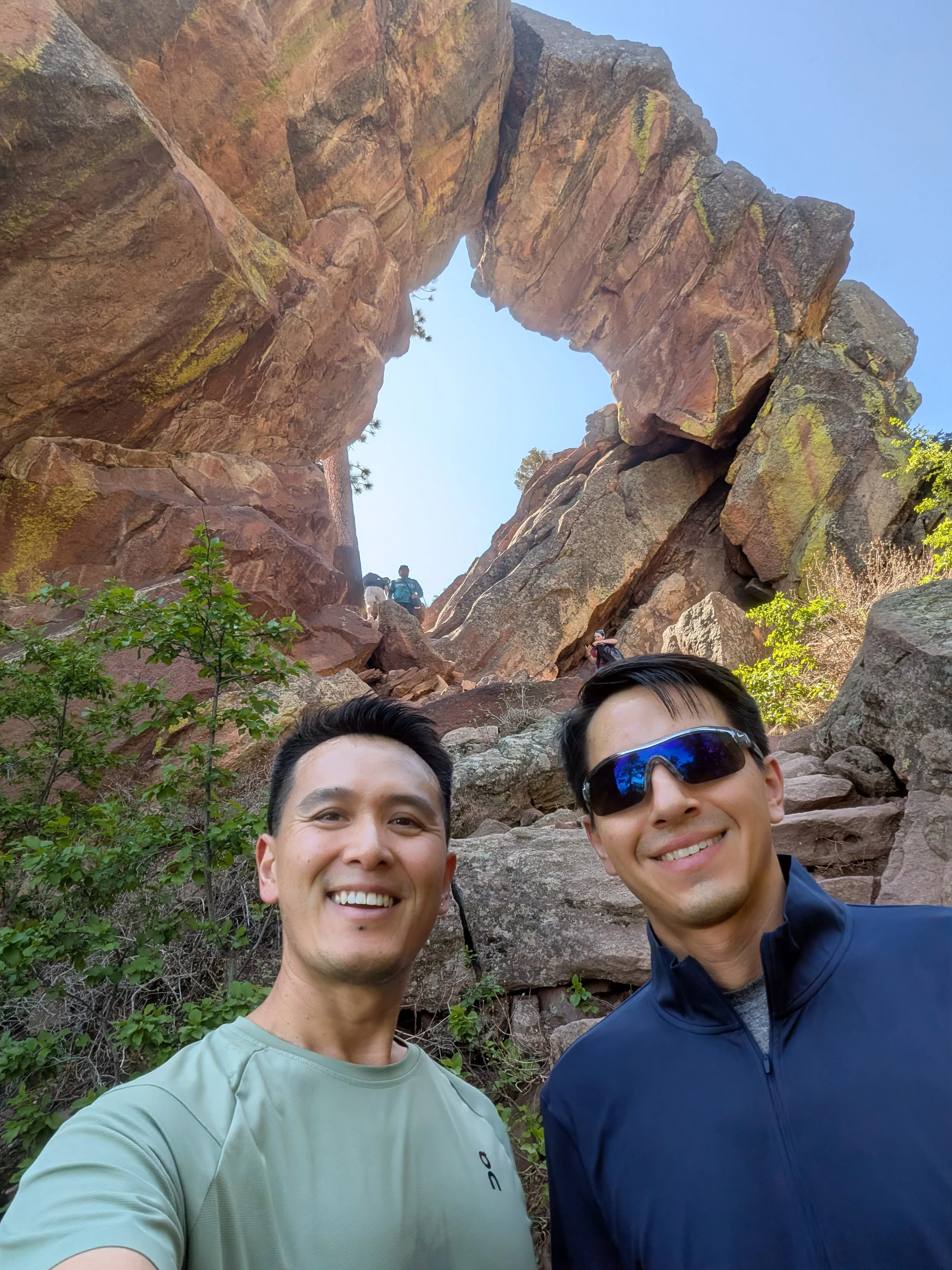 Two smiling men taking a selfie at a rocky mountainous area, under a clear blue sky.