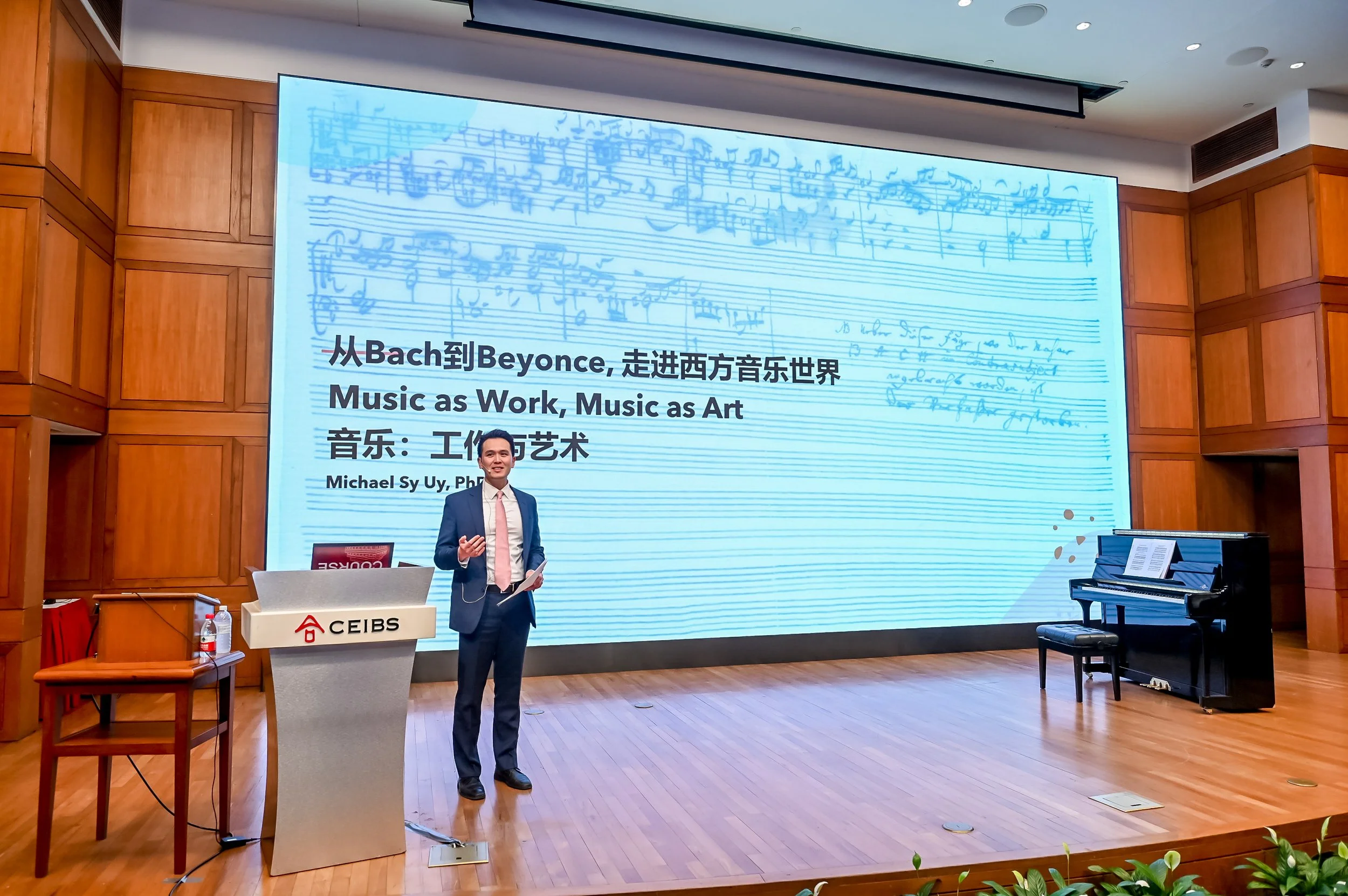 A man in a black suit, white shirt, and pink tie stands on a wooden stage, speaking into a microphone. Behind him, there is a large screen displaying sheet music and Chinese text about Bach and Beyoncé, with the title 'Music as Work, Music as Art' and the speaker's name, Michael Sy Uy, PhD. A grand piano is on the right side, and a lectern with the logo 'CEIBS' is on his left. The background features wood-paneled walls and a ceiling with recessed lighting.