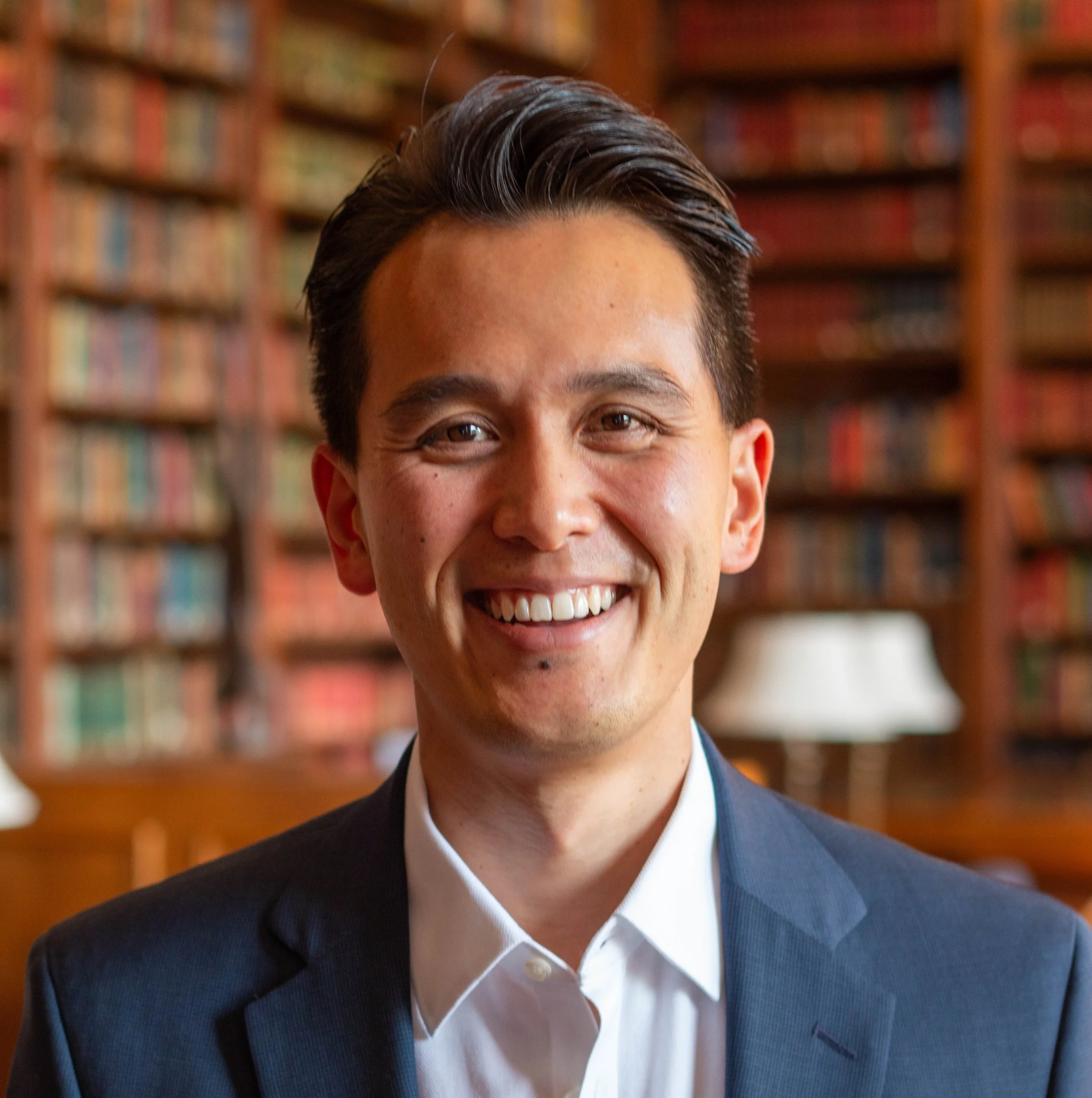 Close-up of a smiling man in a suit with a blurred bookcase in the background.