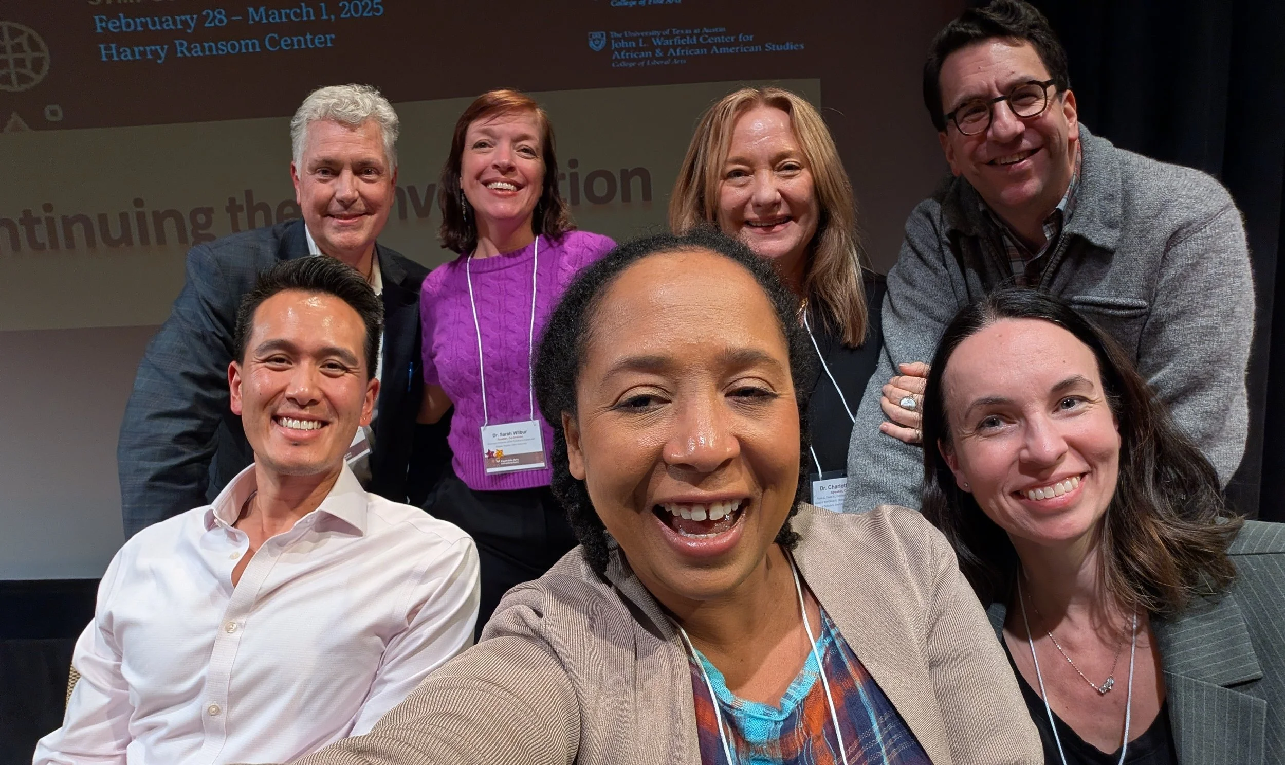 Group of people smiling and taking a selfie at an event. There ar a diverse mix of genders and ethnicities. In the background, there is a projection screen with text and logos, indicating the event dates from February 28 to March 1, 2025, at the Harry Ransom Center, associated with The University of Texas at Austin.