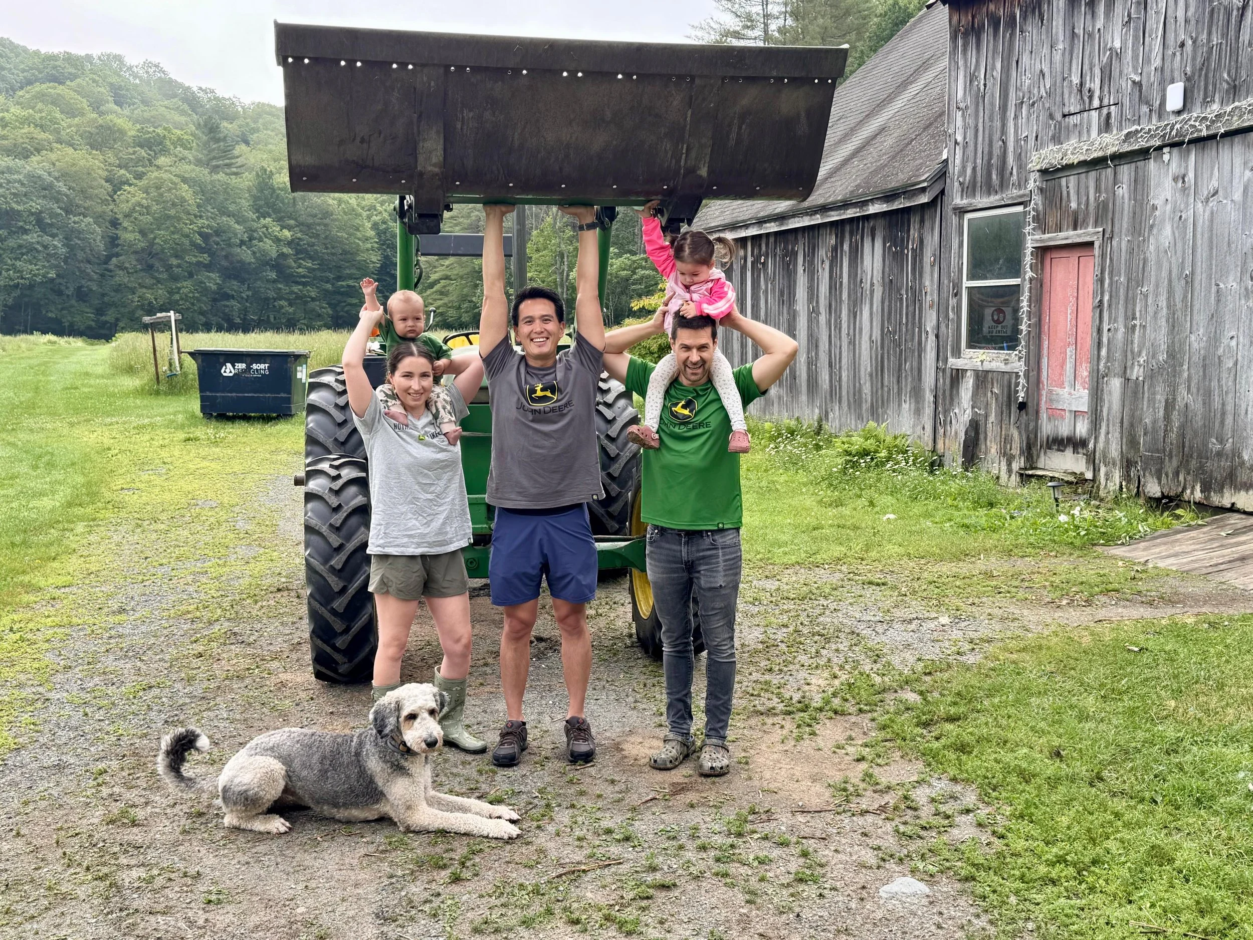 A group of people and a dog posing outdoors in front of a tractor and a rustic wooden building, with green trees in the background. Two children are sitting on and hanging from the shoulders of two adults, who are standing with smiles. The dog is lying on the ground in front of them.