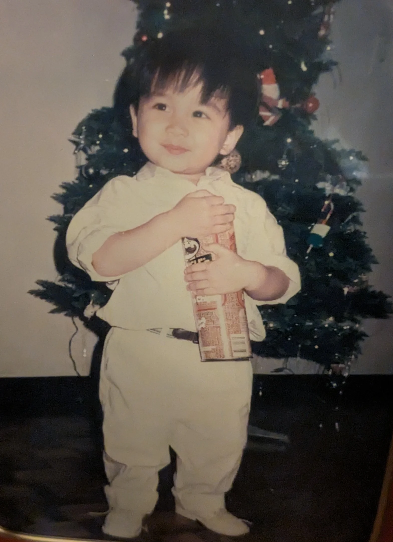 A young child with dark hair, dressed in a white outfit, standing in front of a decorated Christmas tree, holding a can of Pringles.