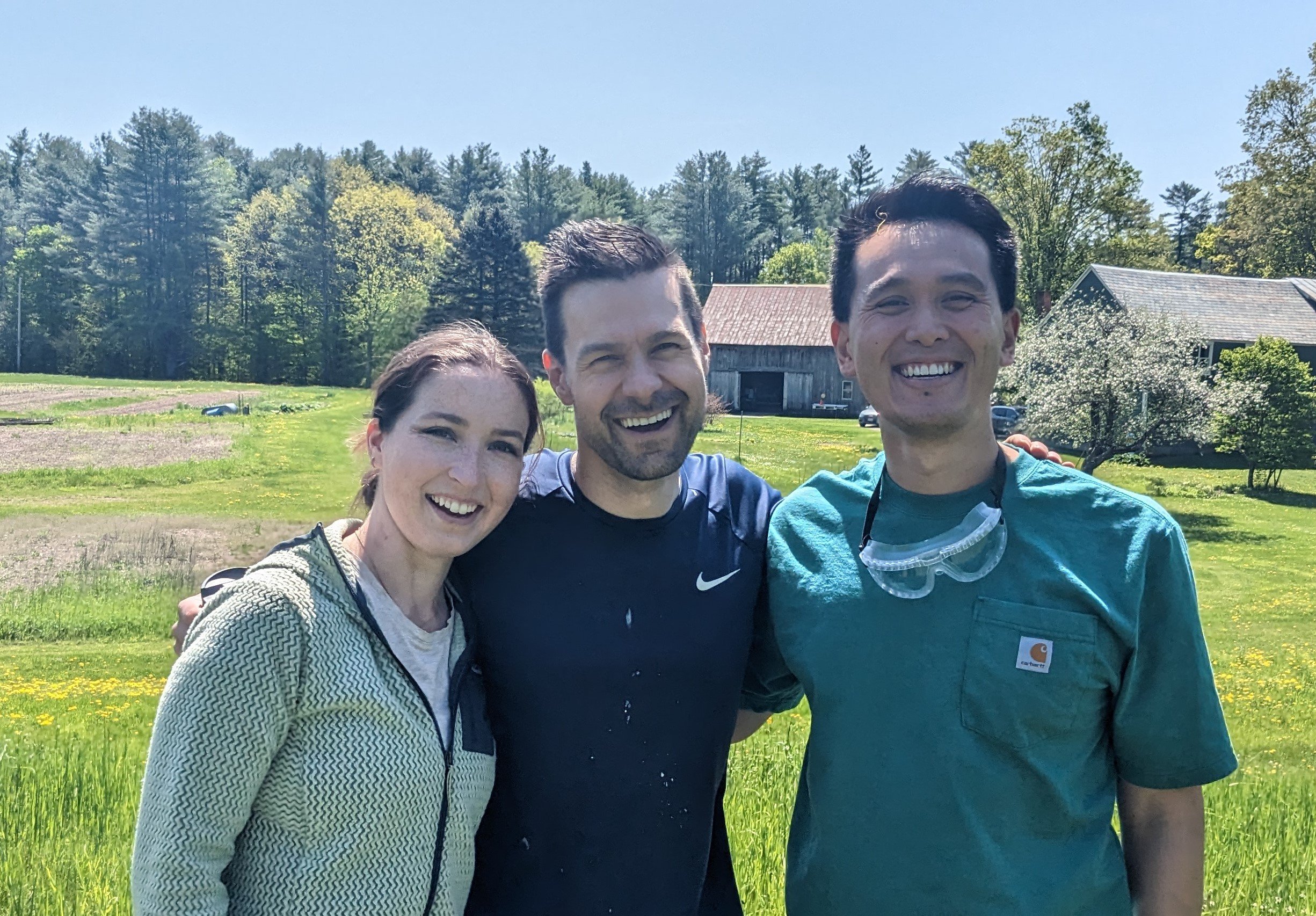 Three smiling people standing outdoors on a sunny day, with trees and farm buildings in the background.