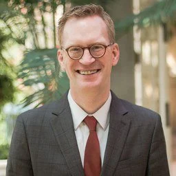 Portrait of a smiling man wearing glasses, a dark blazer, white shirt, and red tie outdoors with trees in the background.