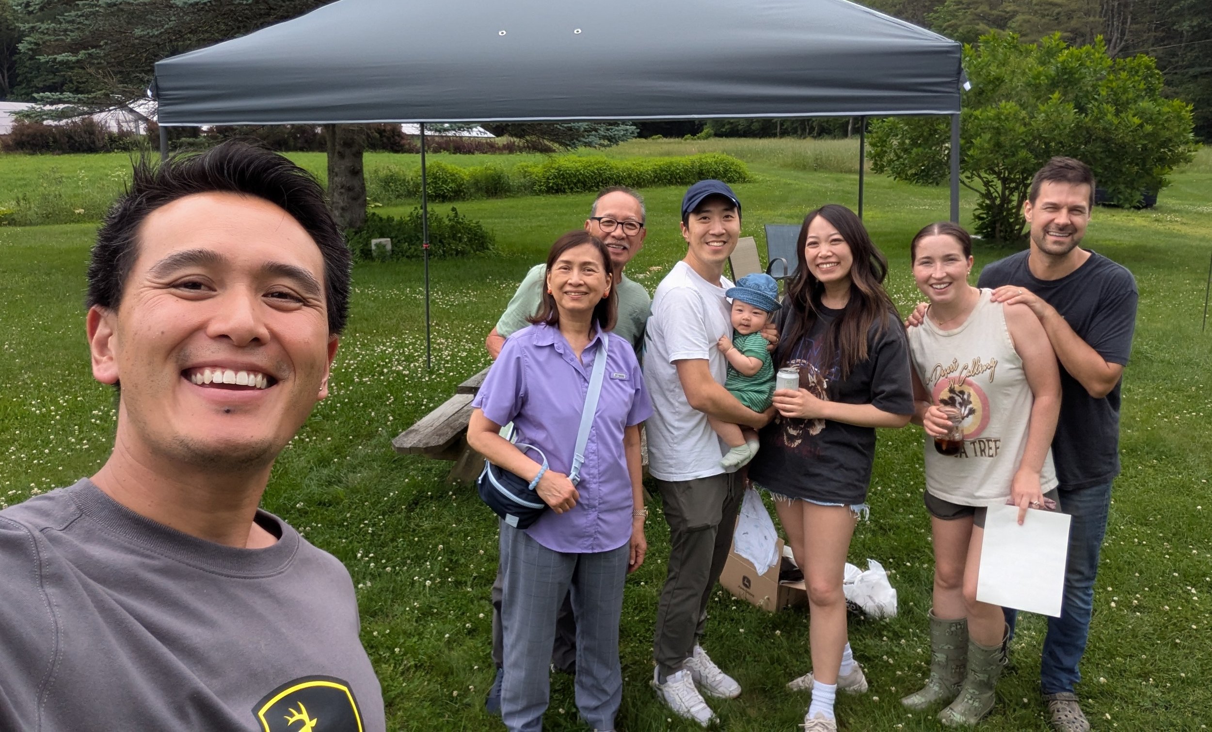 A group of eight people, including one baby, is gathered outdoors on a grassy area under a canopy tent, smiling for a photo, with trees and greenery in the background.