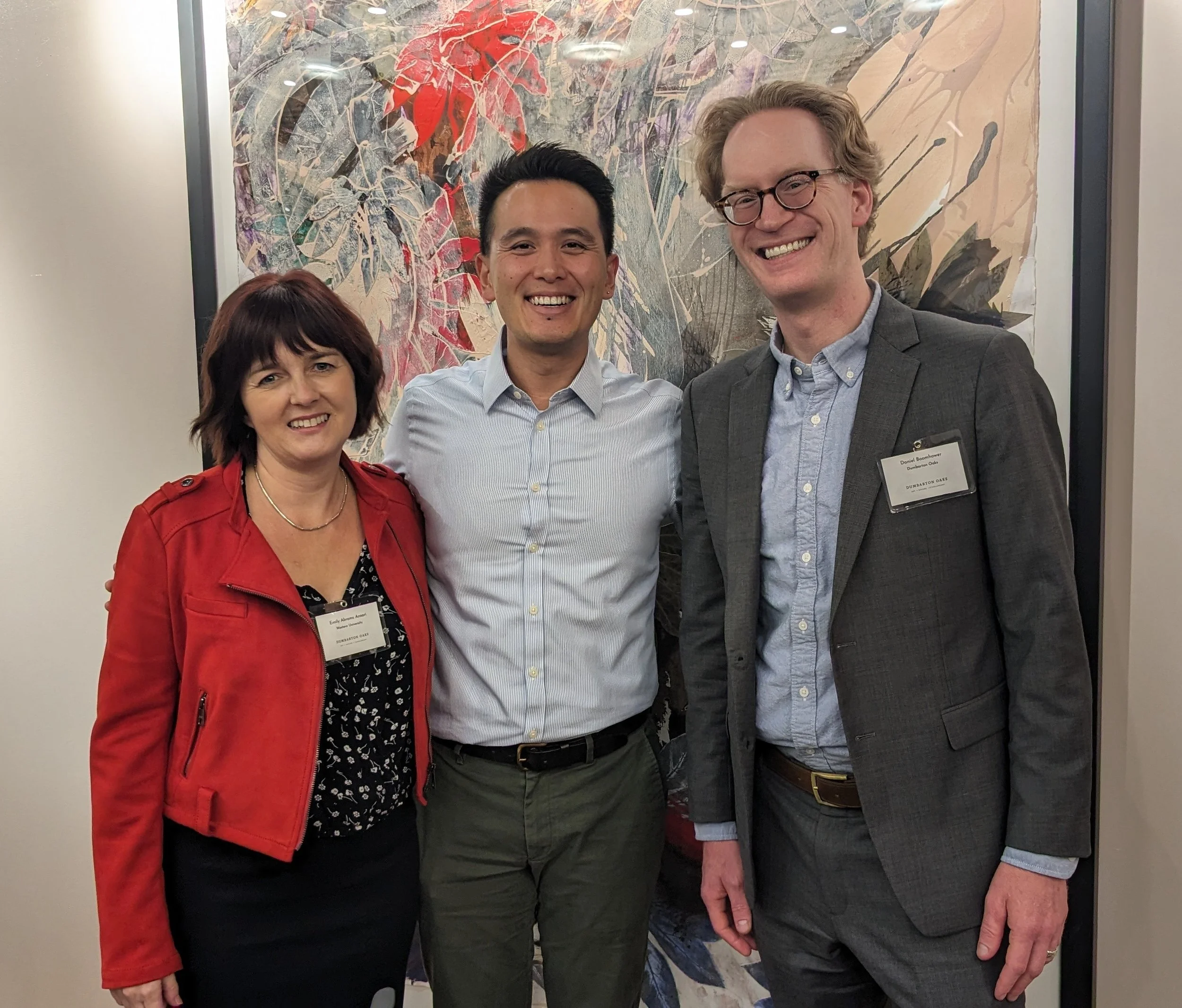 Three people are standing together indoors, smiling at the camera. Behind them is abstract wall art with various colors and textures.