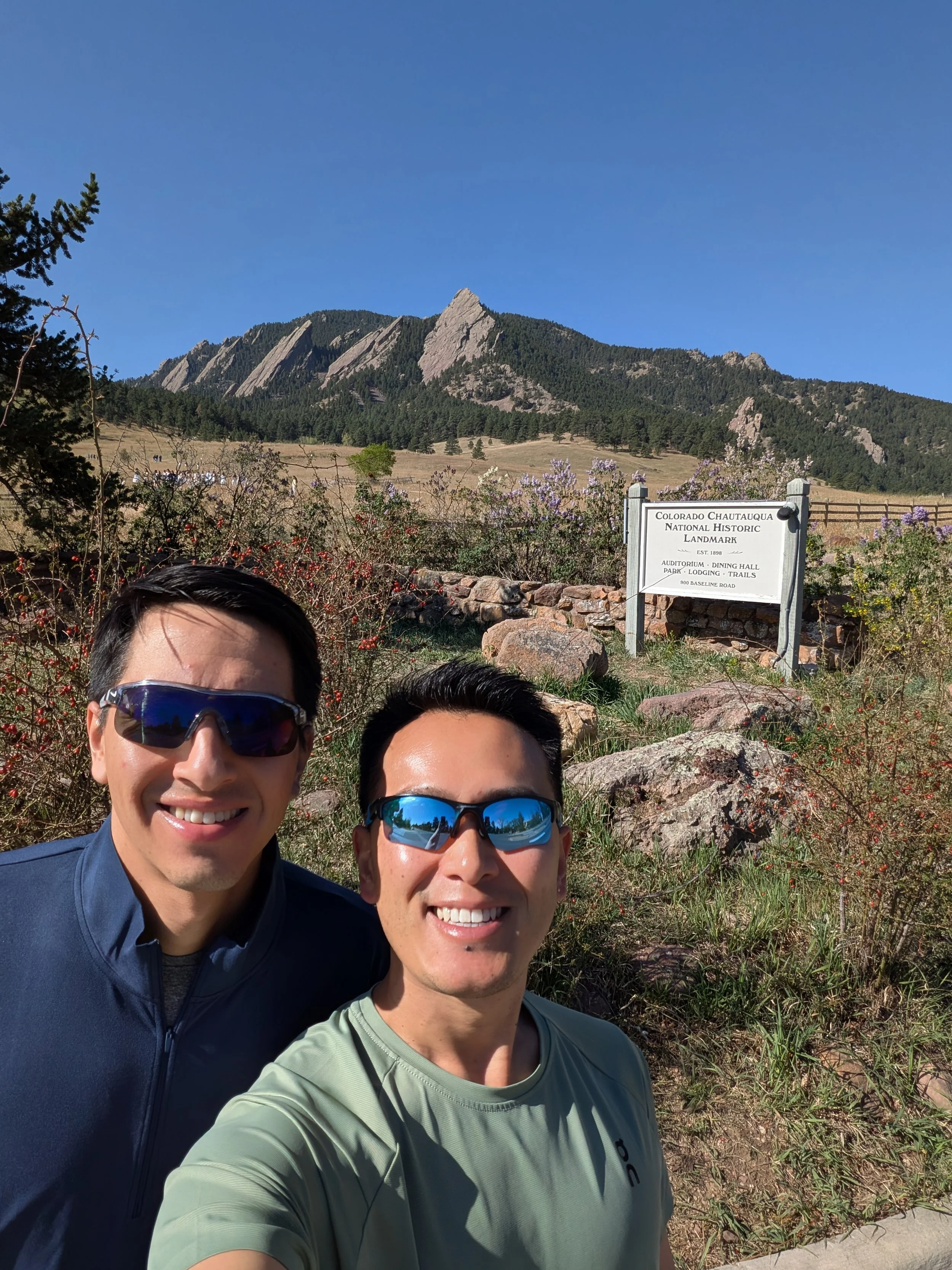 Two men wearing sunglasses taking a selfie in front of a mountain landscape with a sign that reads "Colorado Chautauqua National Historic Landmark."
