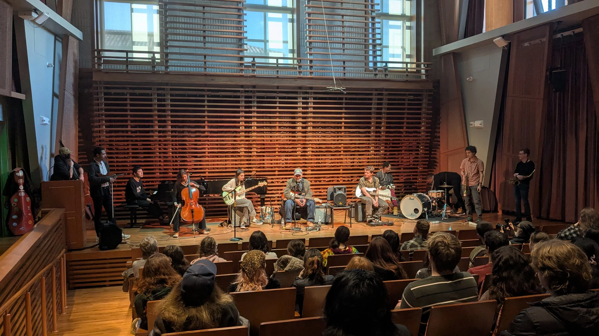 A musical performance on a stage with a band of seven musicians, including guitarists, a bassist, a drummer, and keyboard players, in front of an audience in an auditorium with wooden wall accents and large windows.