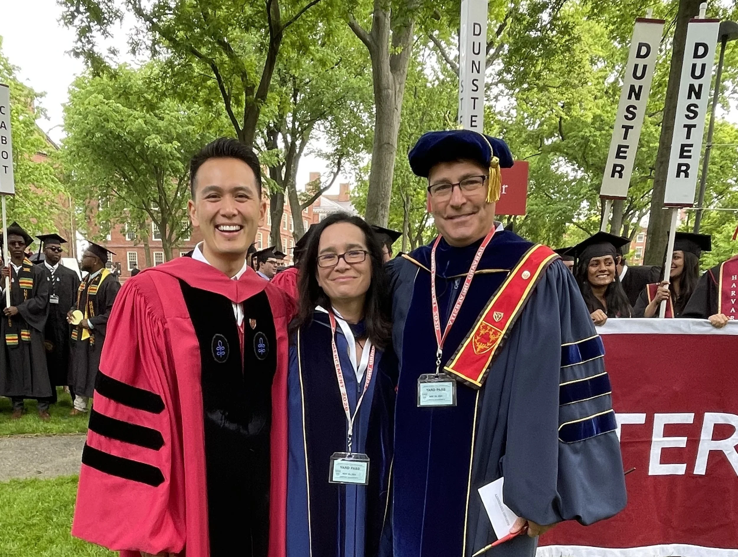 Three graduates in academic regalia smiling at an outdoor graduation ceremony with trees and signs in the background.