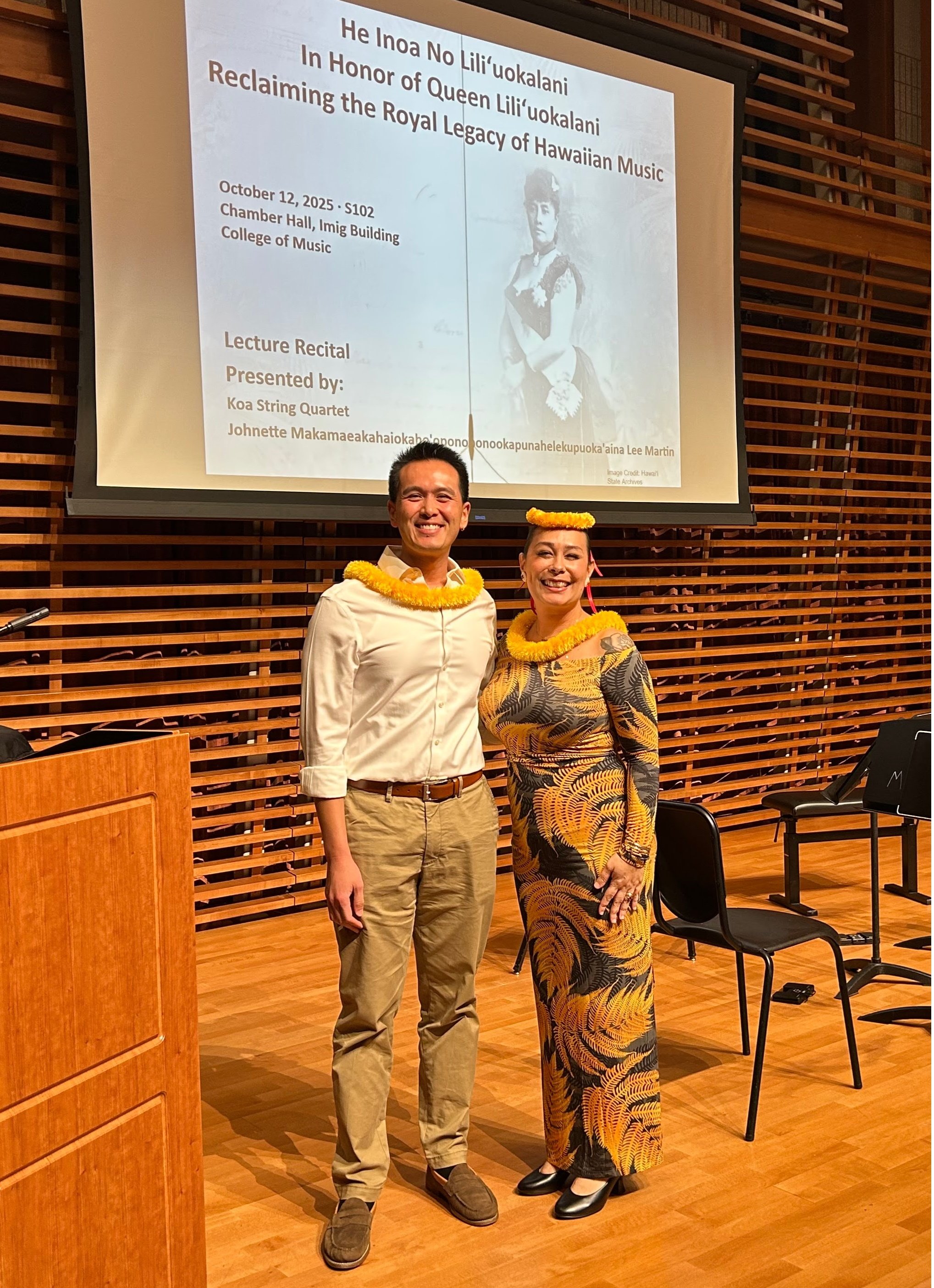 Two individuals standing in front of an auditorium with a large screen displaying an event announcement. The person on the left is a man in a white shirt and beige pants, smiling, with a yellow lei around his neck. The person on the right is a woman in a patterned dress, also smiling, with a yellow leis around her neck and a flower headpiece. The event details on the screen mention a lecture recital about Hawaiian music, scheduled for October 12, 2025, at the College of Music.