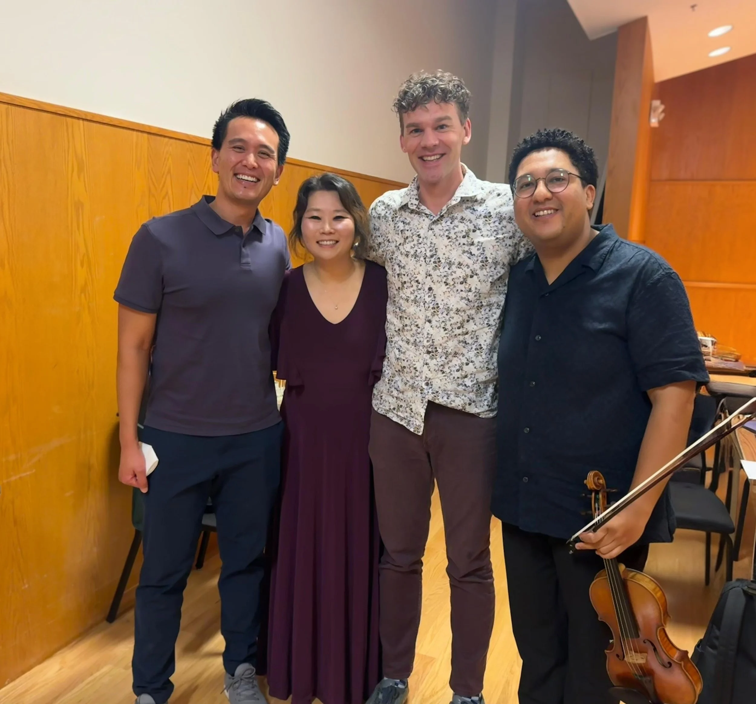 Four people smiling together in a room, with a woman in the middle and three men around her, one holding a violin. The background features wooden paneling and a music stand with sheet music.