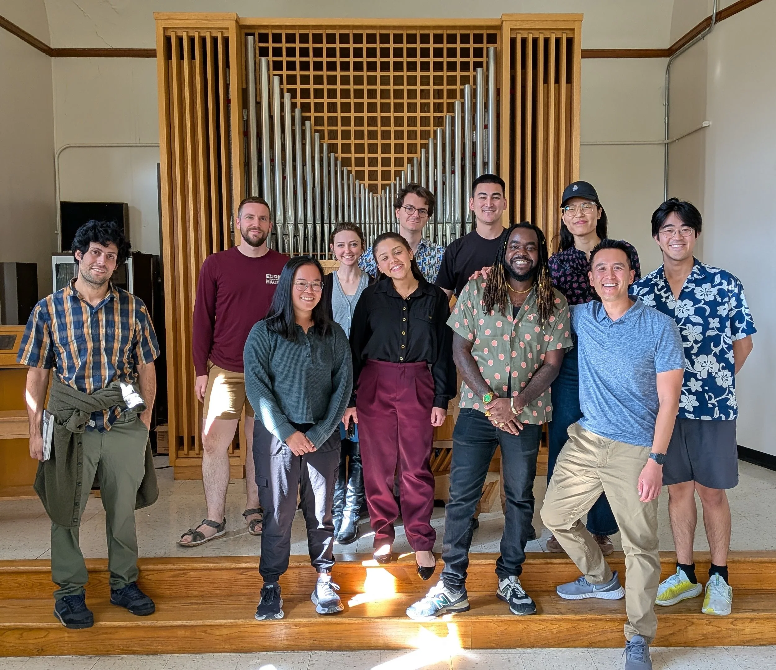A diverse group of people standing together and smiling in front of a large pipe organ against a wooden wall inside a building.