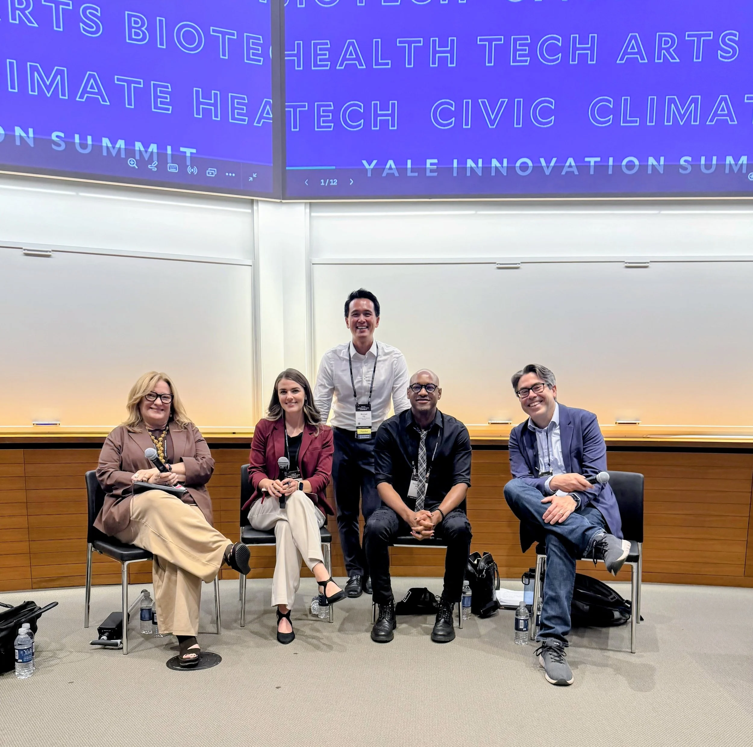 Group photo of five diverse people, four sitting and one standing, at a conference or panel discussion, with water bottles and bags on the floor, in front of a large screen displaying a colorful presentation slide for Yale Innovation Summit.