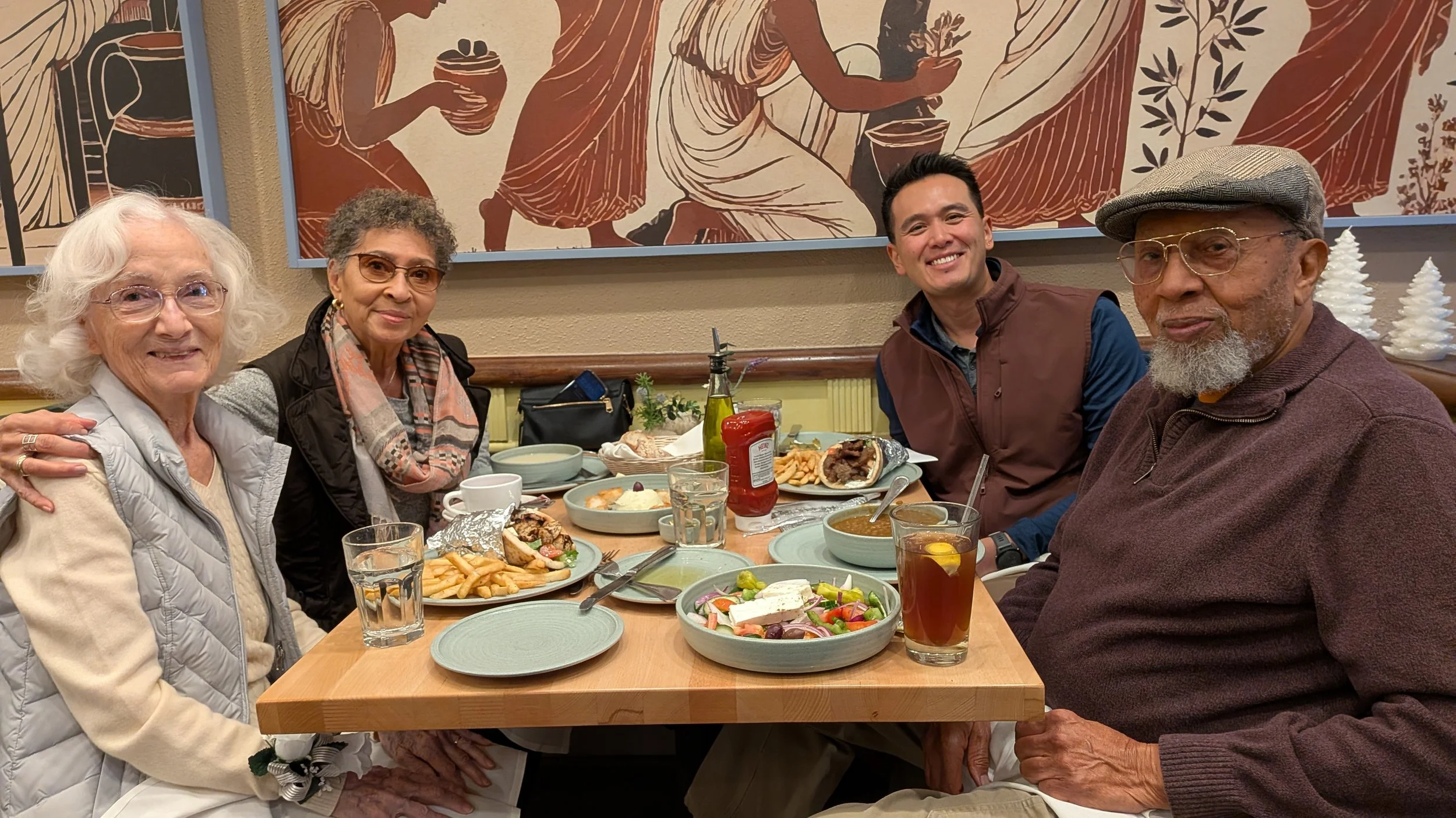 A group of four people sitting around a table with various foods. They are smiling and enjoying a meal at a restaurant. There are plates of salad, fries, sandwiches, and drinks on the table, with a decorative mural on the wall behind them.