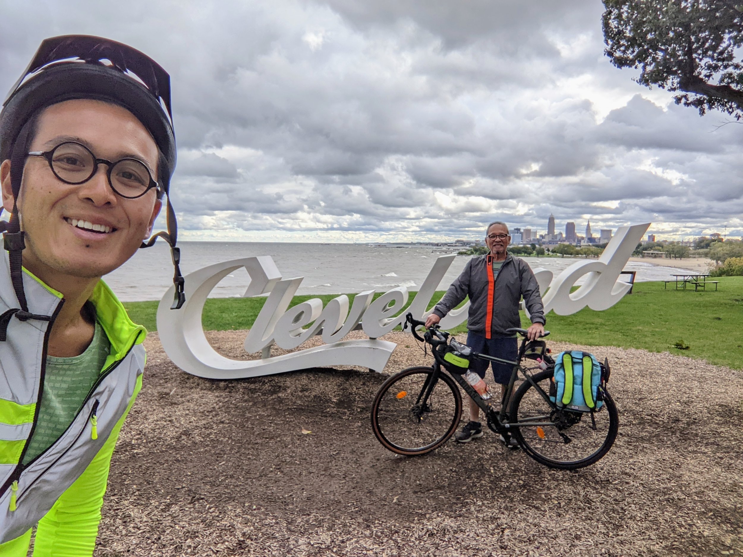 Two men with a bicycle standing near a large white 'Cleveland' sign on a grassy area by water with city skyline in the background, cloudy sky overhead.
