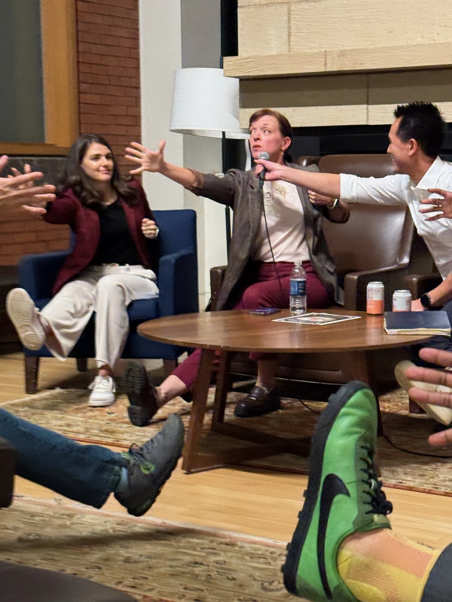 A group of people seated on a carpet and chairs in a room, engaging with each other. One person, holding a microphone, appears to be speaking or presenting. Others are listening and smiling, with some extending their hands toward the speaker. The scene suggests a casual or informal gathering, possibly a discussion or talk.