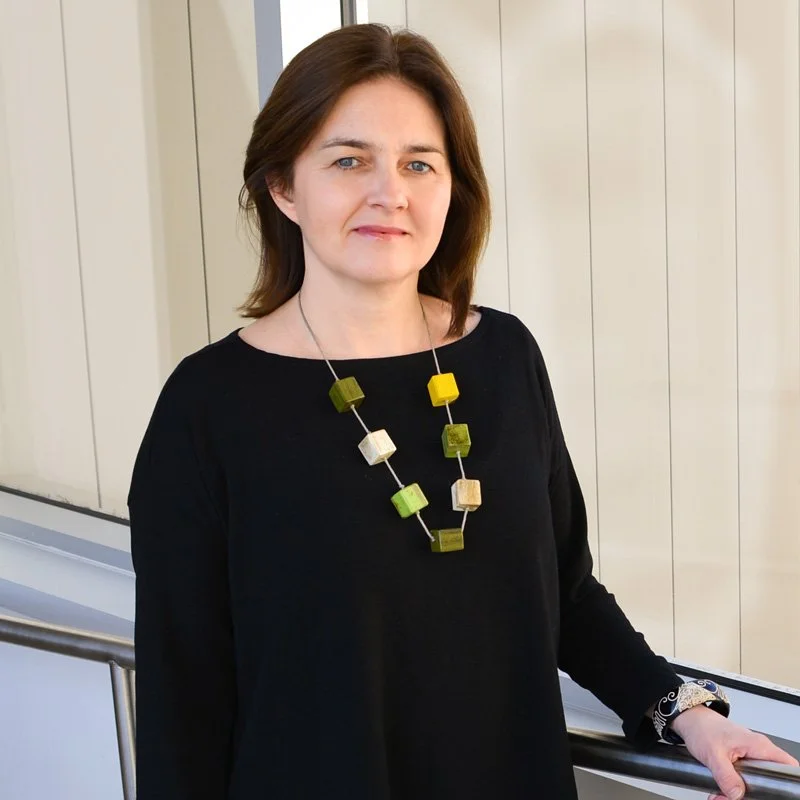 A woman with shoulder-length brown hair standing indoors near a window with light-colored vertical blinds. She is wearing a black long-sleeve top and a colorful cube-shaped beaded necklace.