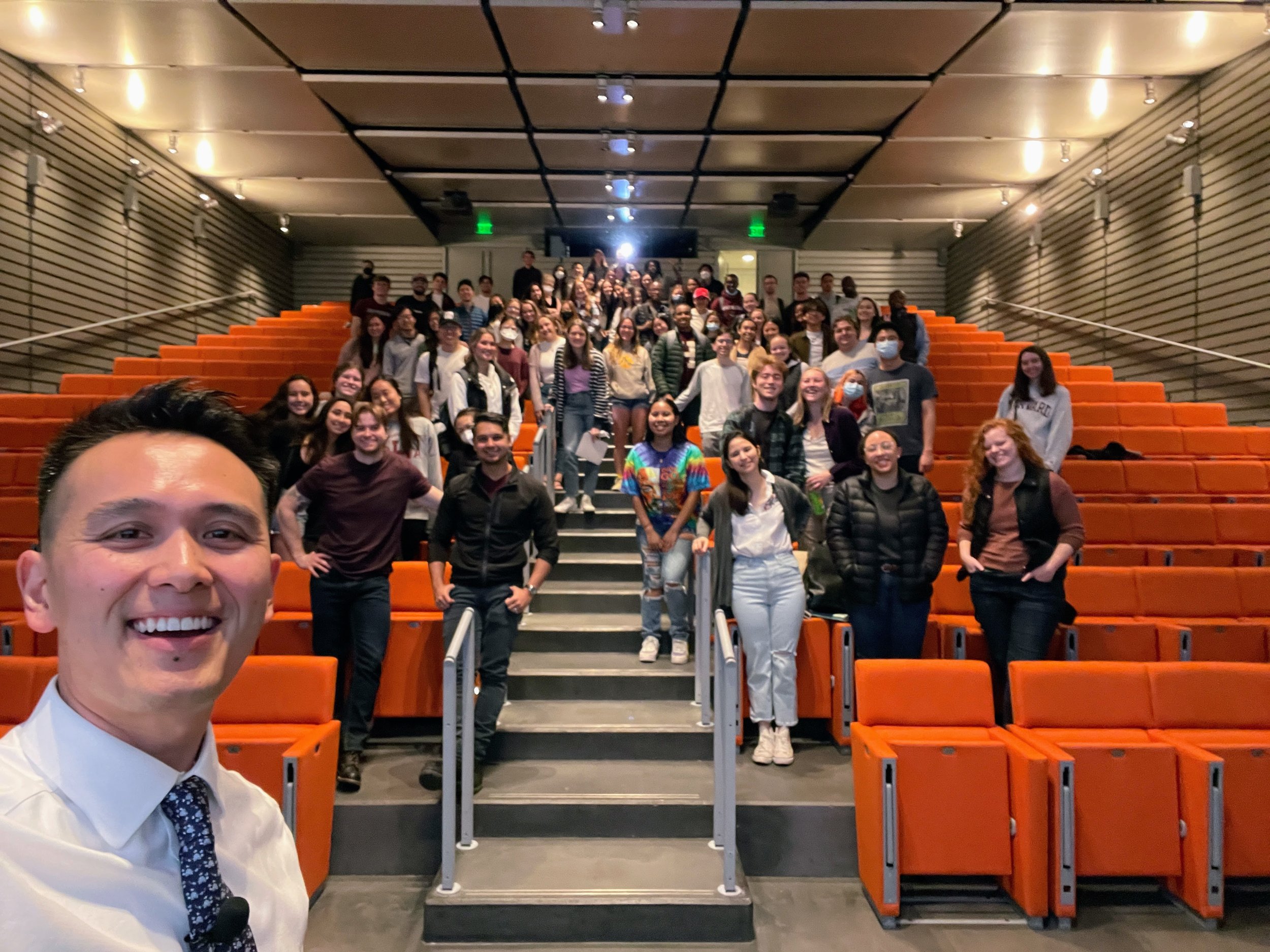 A selfie of a man in a white shirt and patterned tie, smiling, with a large group of people standing on orange theater seats and stairs behind him in a theater lobby.